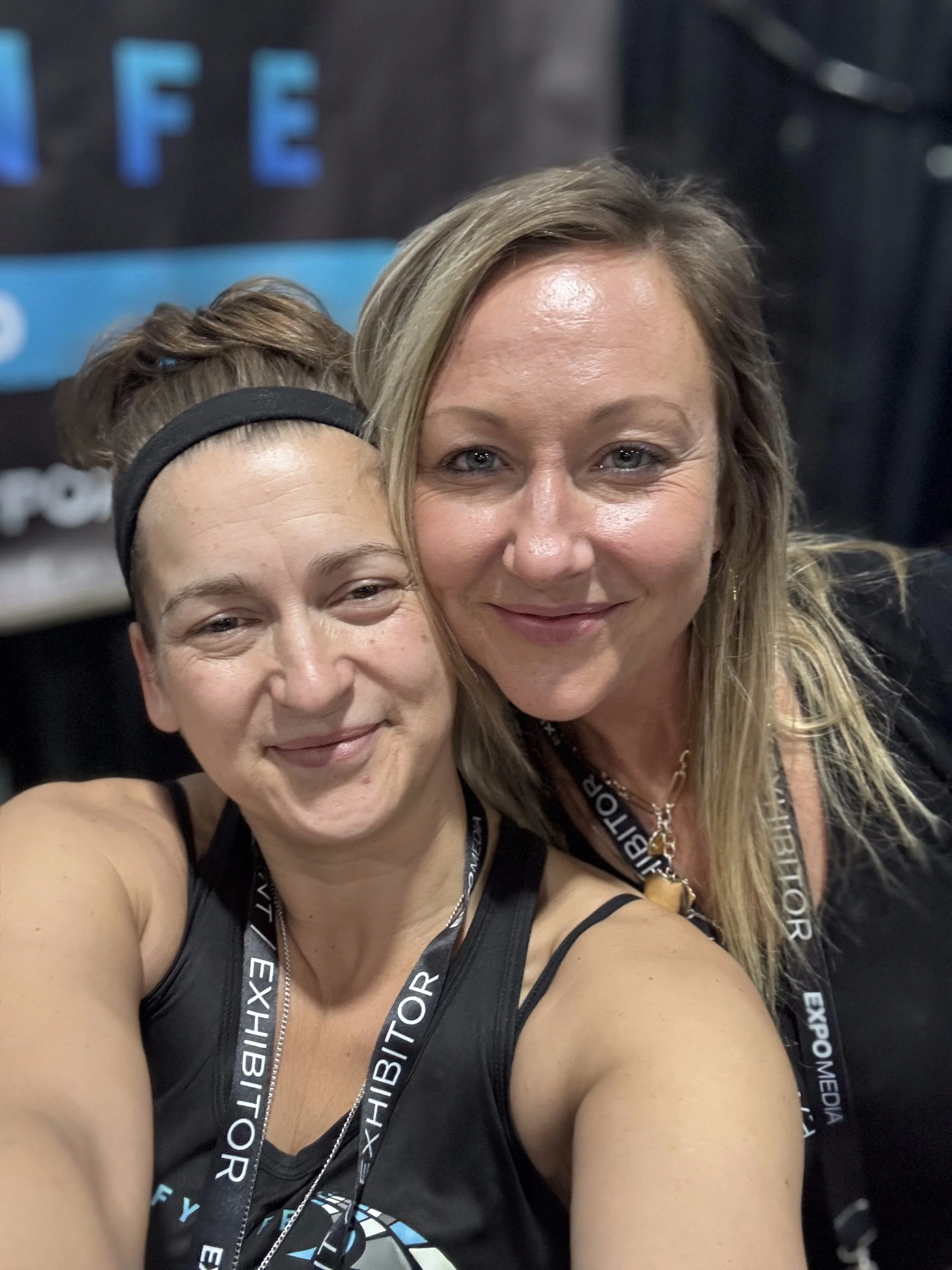 Two women taking a selfie at an expositor event, smiling. One woman has dark hair tied back with a black headband, wearing a black tank top and exhibitor lanyard. The other woman has blonde hair, wearing a black outfit and lanyard, with a background of a sign and dark curtains.