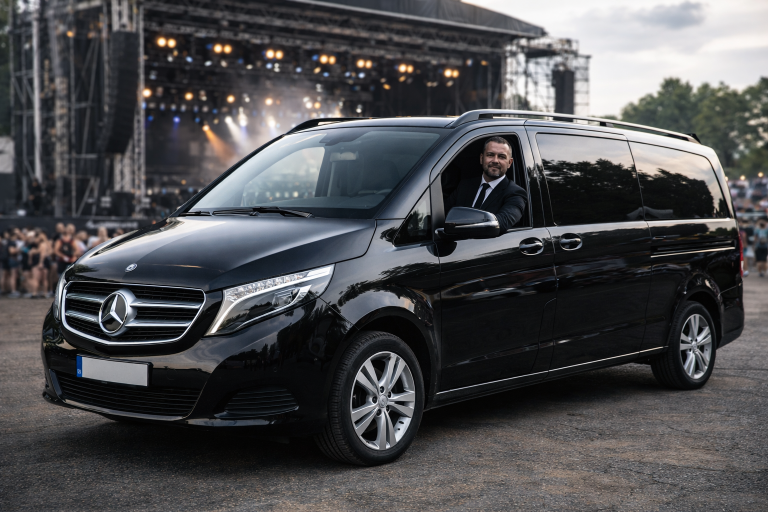 Conductor masculino en suit negro sentado en un vehículo negro Mercedes-Benz en un concierto con escenario y público de fondo.