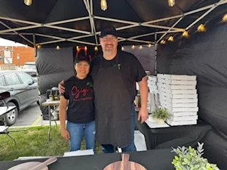 Two people standing under a canopy tent at an outdoor event, with a table in front of them and stacked pizza boxes in the background.