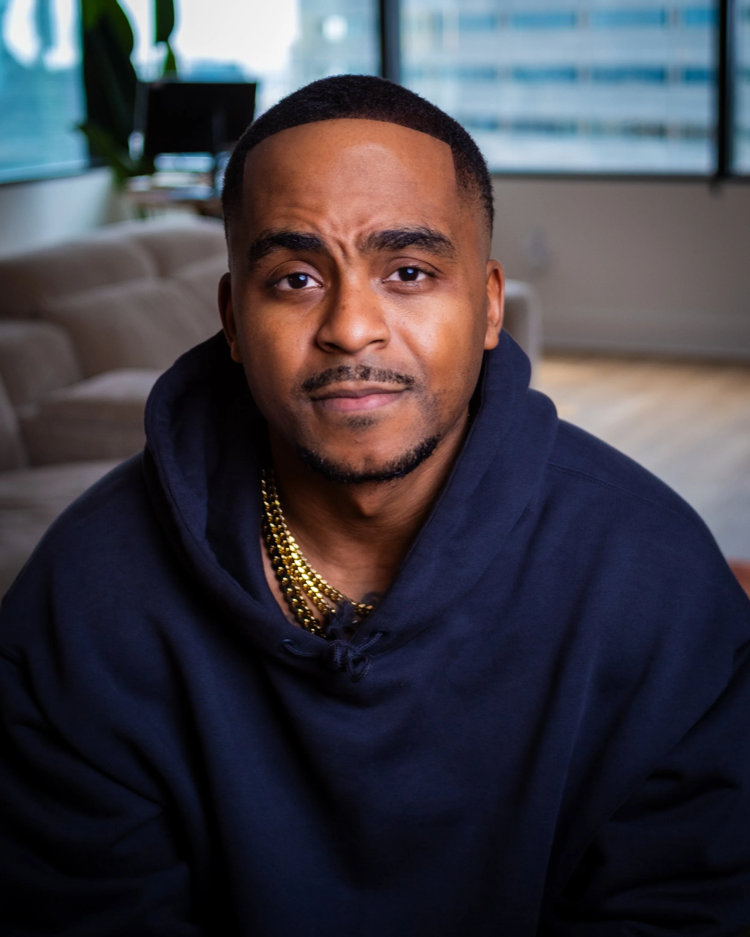A man with short black hair, dark eyebrows, and a goatee, looking at the camera with a neutral expression. He is wearing a black hoodie and a gold chain. The background shows an office or living room with large windows and a sofa.