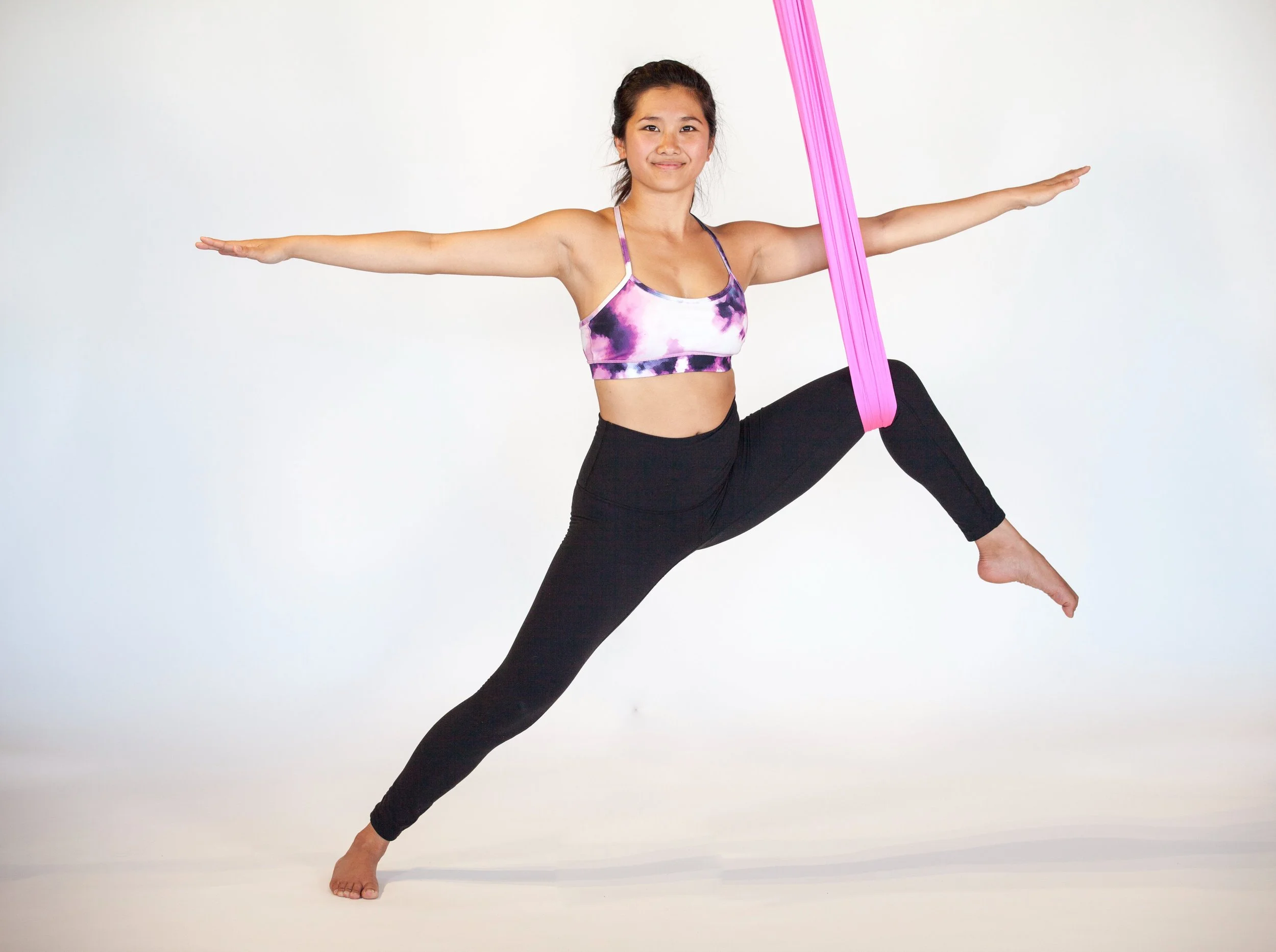 Young woman practicing aerial yoga, standing on one leg in a yoga pose, with arms extended horizontally, wearing a purple and white sports bra and black leggings, against a plain white background.
