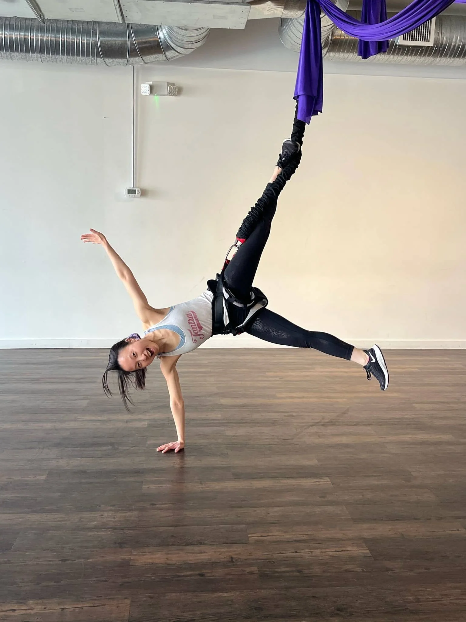 A woman in athletic clothing performing a one-handed yoga pose on a wooden floor in a studio with ductwork and a purple aerial silk hanging from the ceiling.