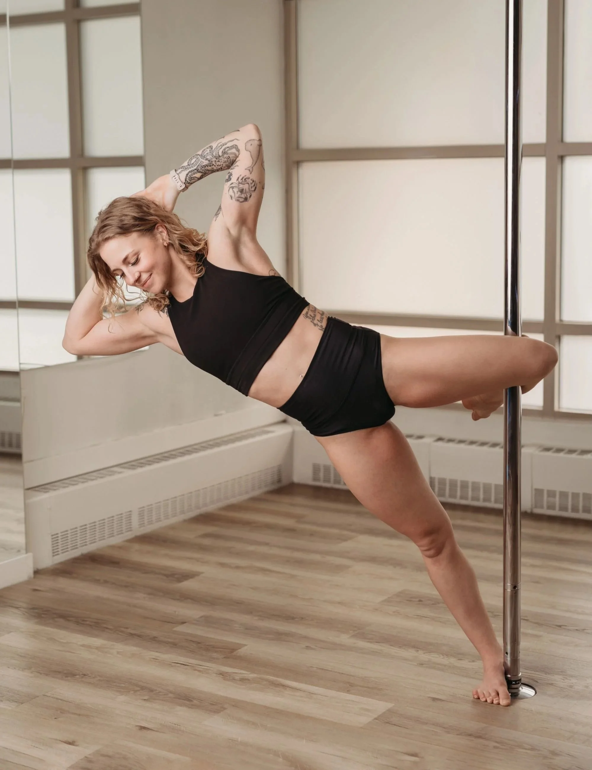A woman with tattoos practicing pole dancing in a studio with wooden floors and white paneled walls.