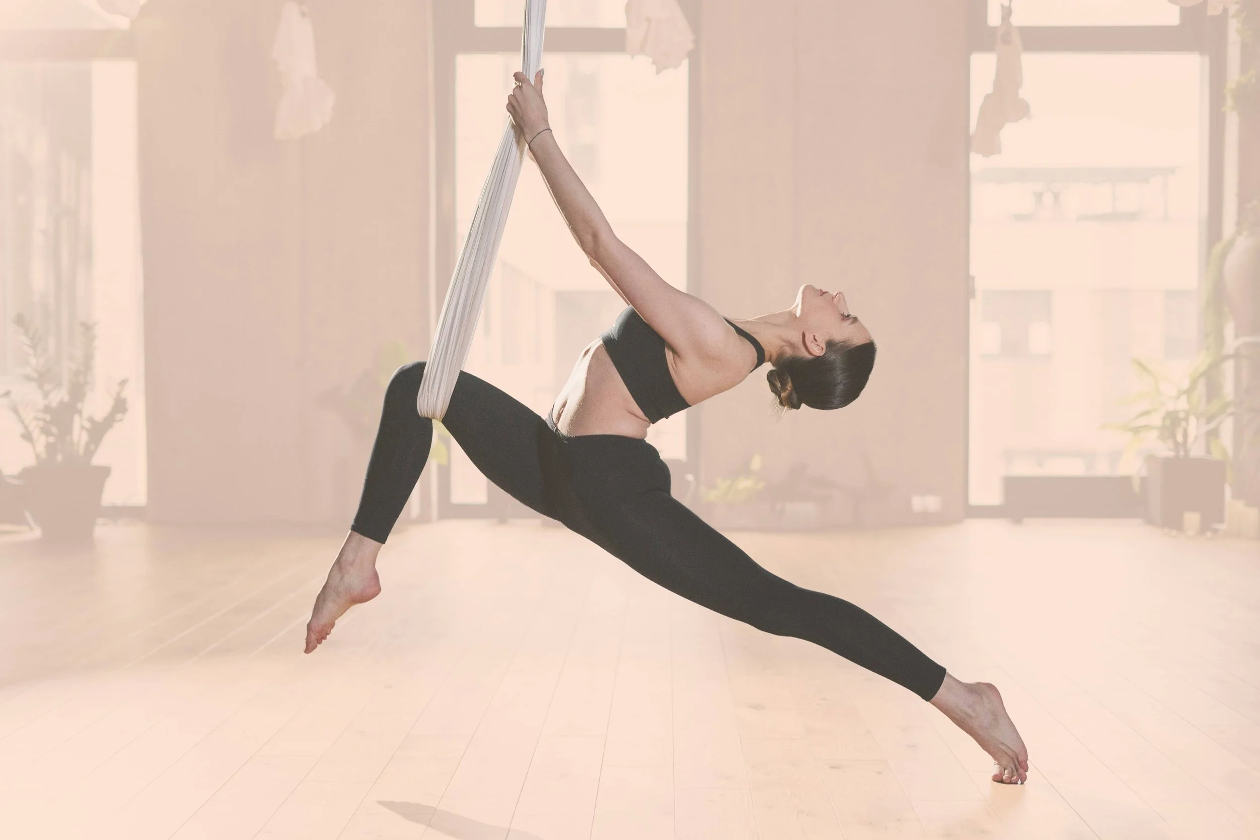 A woman practicing aerial yoga in a brightly lit room with large windows, hanging from a white fabric sling while balancing on the floor.