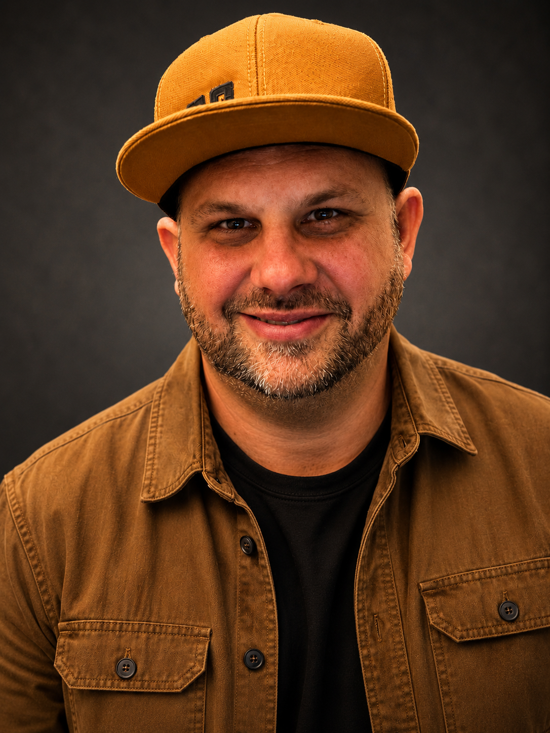 A man with a beard and mustache wearing a tan baseball cap, brown jacket, and black shirt, smiling against a dark gray background.