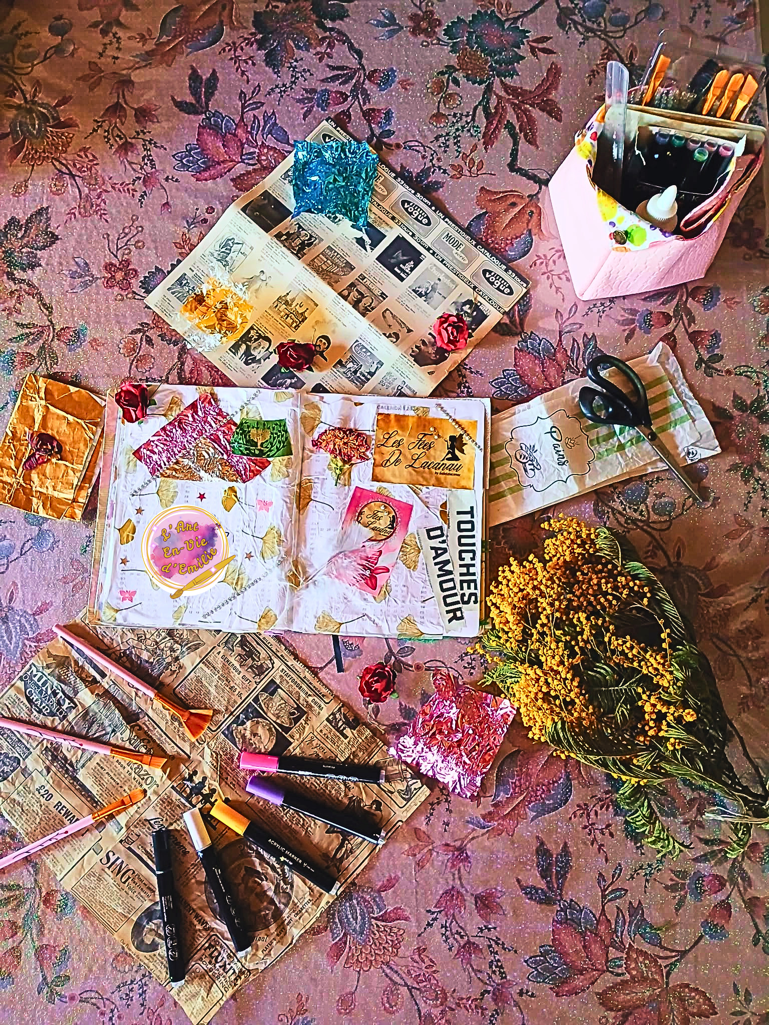 Les objets pour faire de l'artisanat ou de la décoration, posés sur un tapis à motifs floraux. Il y a des flowers, des marqueurs, des pots de peinture, des magazines, et des papiers colorés.