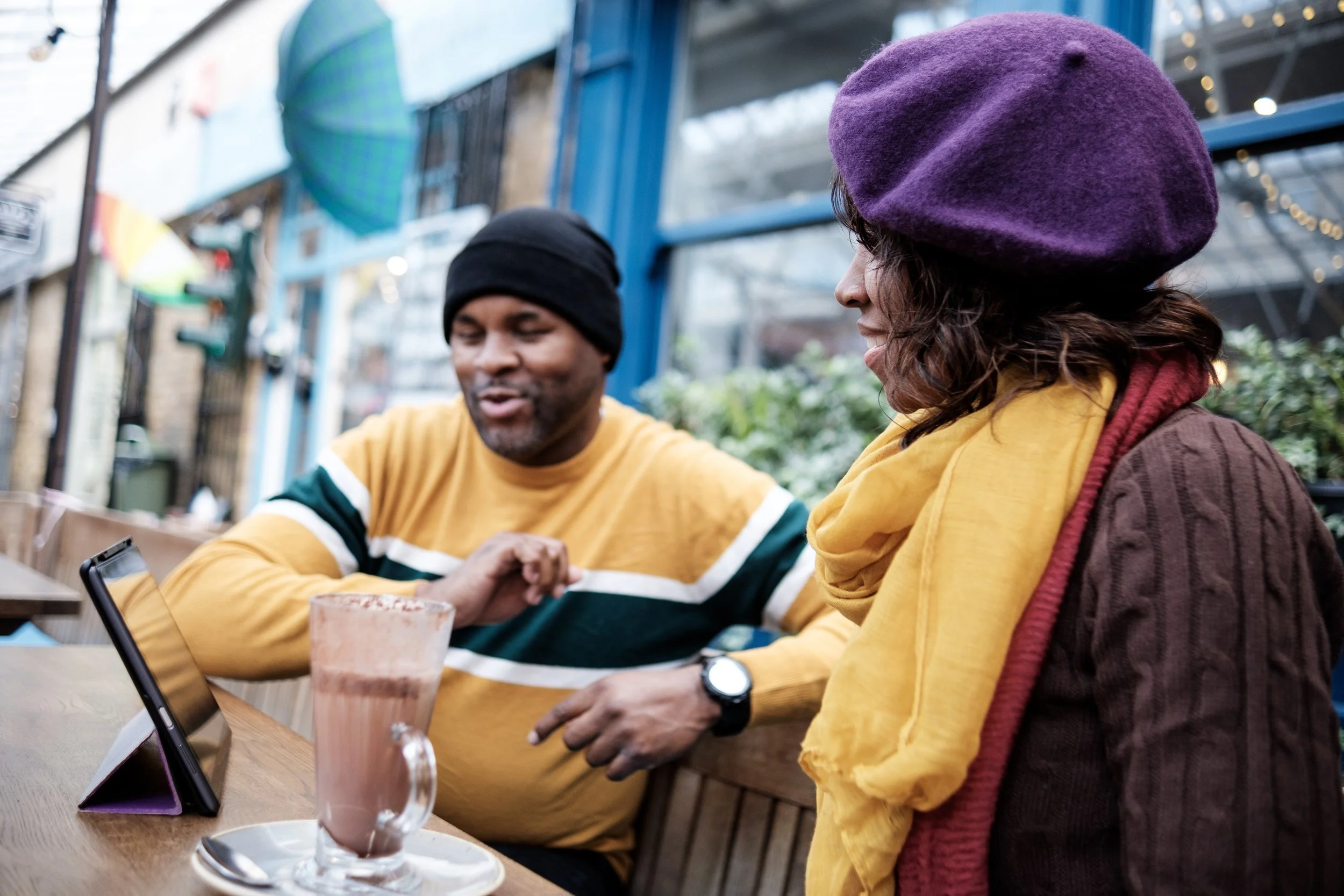 A man and woman sitting at an outdoor table, looking at a tablet device, with a tall glass of chocolate milkshake in front of the man. The woman is wearing a purple beret and yellow scarf, and the man is wearing a black beanie and a yellow, green, and white striped sweater.