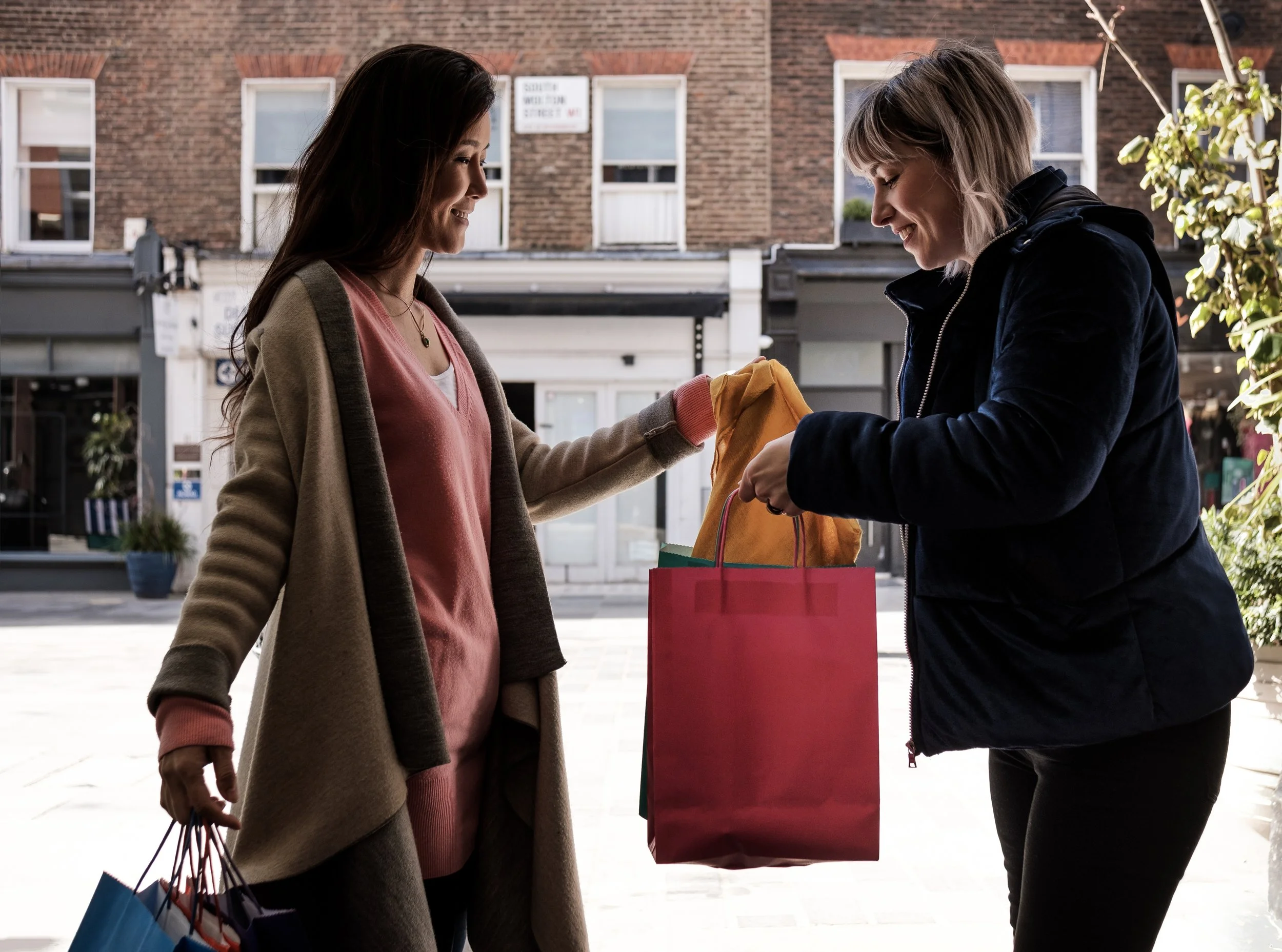 Two women exchanging shopping bags on a city street.