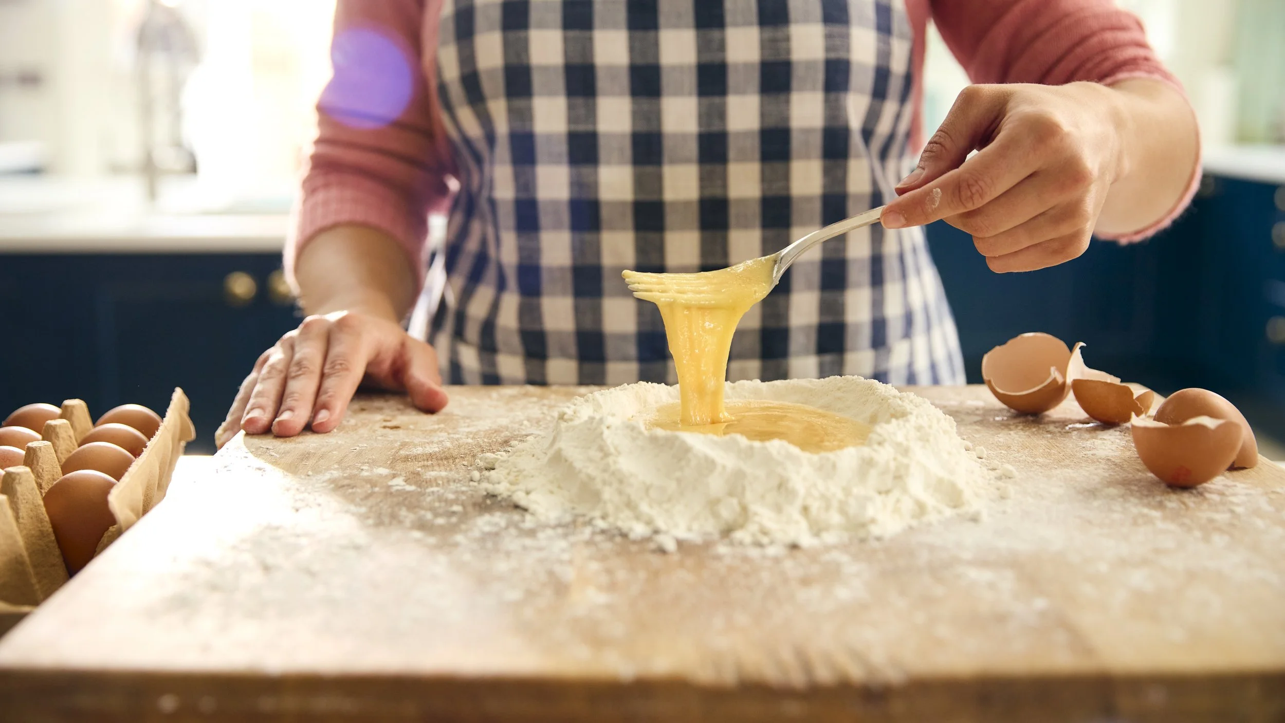 Person mixing eggs and flour in a kitchen with eggshells and a carton of eggs on the table.