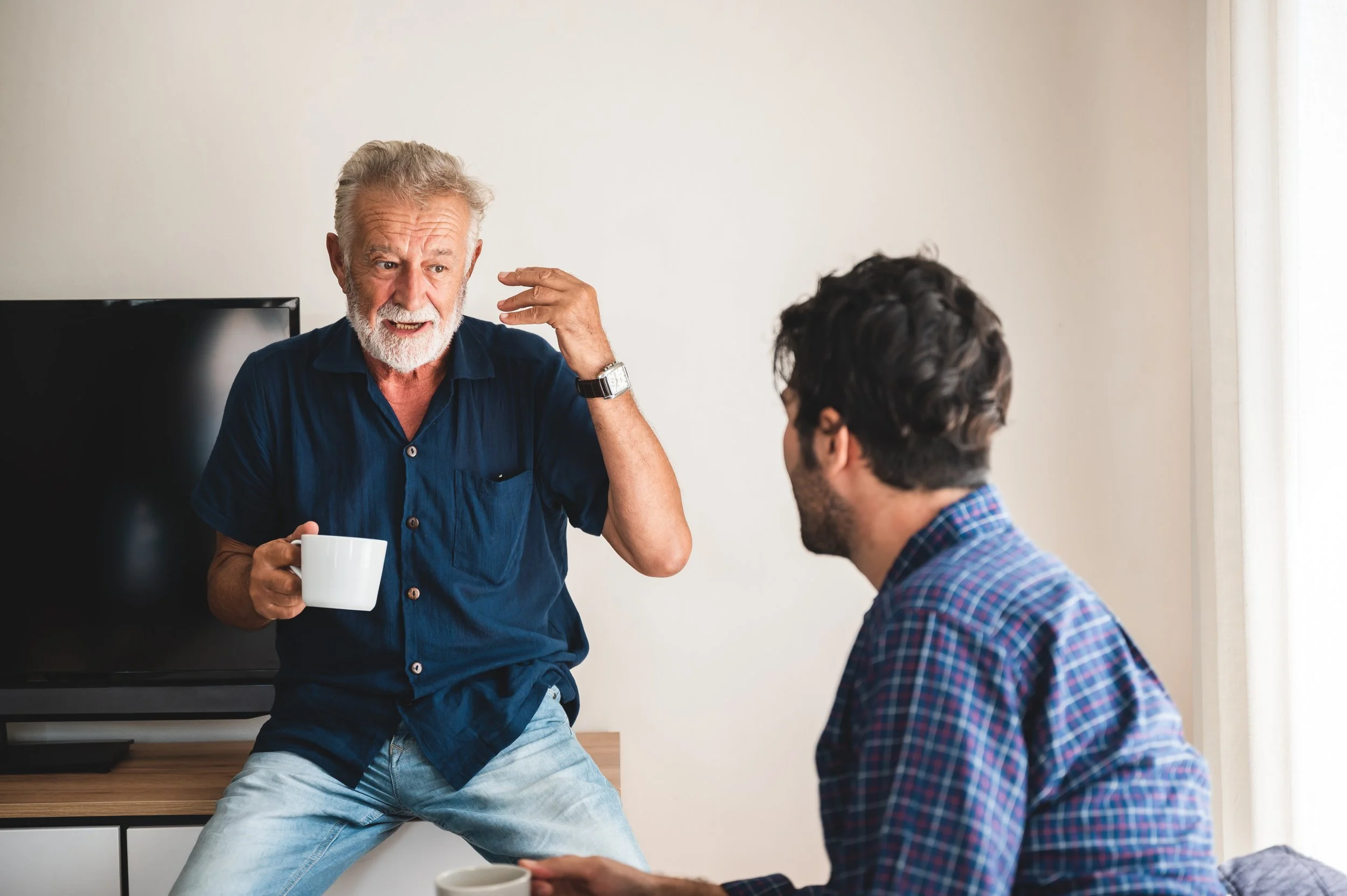An older man with a white beard and gray hair standing, gesturing while talking to a younger man sitting on a couch, holding a mug, in a living room.