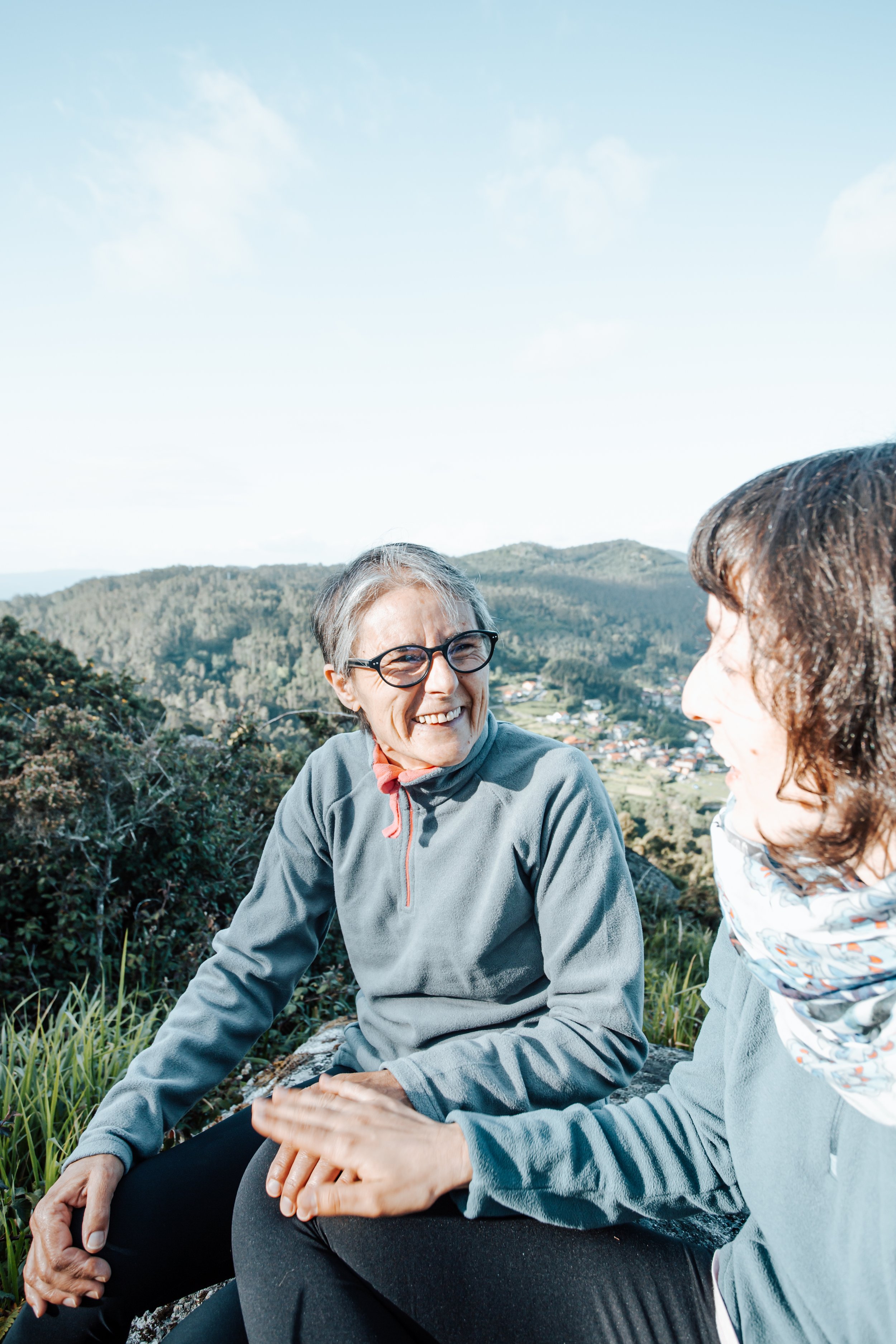 Two women sitting outdoors on a hillside, smiling at each other with a scenic landscape in the background, including hills, trees, and a clear blue sky.