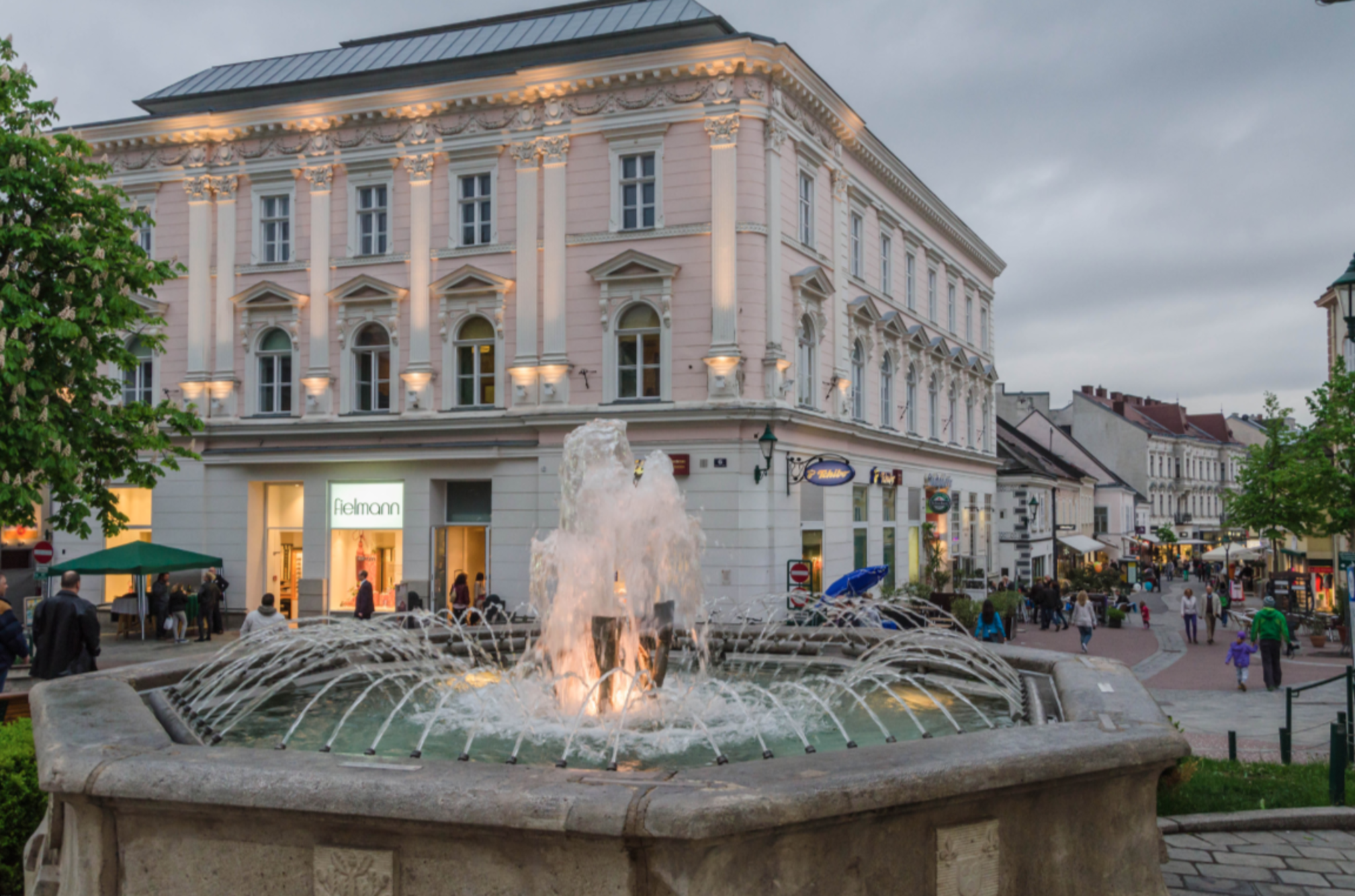 Blick auf einen Springbrunnen in der belebten Mödlinger Fußgängerzone mit historischen Gebäuden und Menschen, die spazieren gehen.