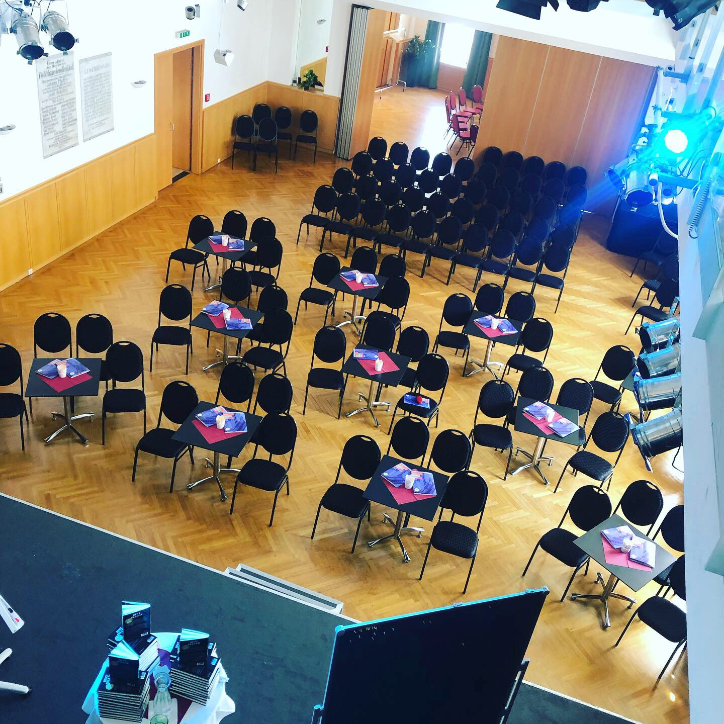 Ein leeres Veranstaltungsraum mit schwarzen Stühlen und Tischen, die mit blauen und roten Tischsets und kleinen Verpackungen bedeckt sind. Es ist für eine Veranstaltung vorbereitet, mit einem Blick auf die Bühne im Vordergrund.