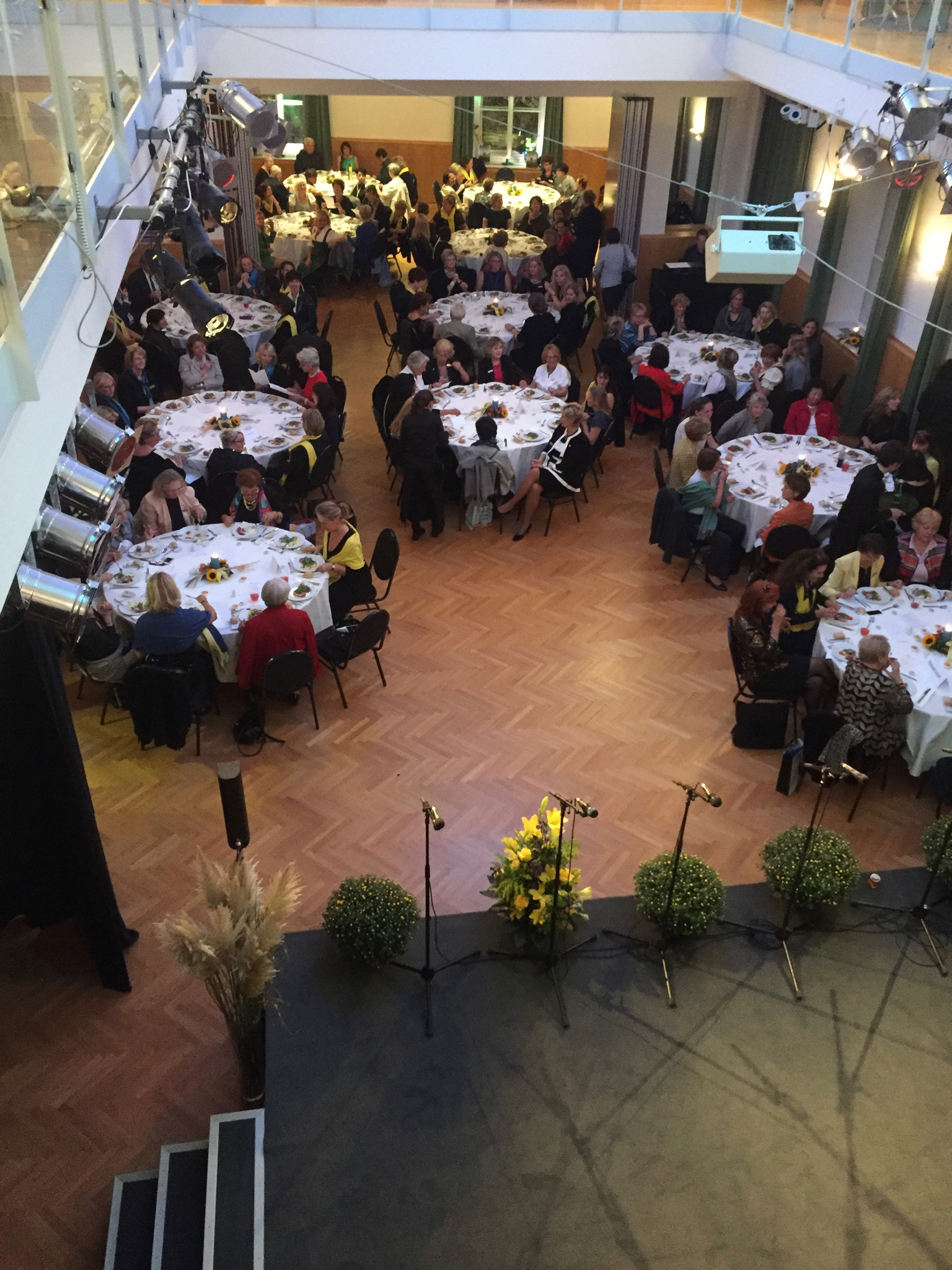 Festsaal mit festlich gedeckten Tischen, ein Bühnenbereich mit Mikrofonen und Pflanzen, voll mit Gästen bei einem formellen Event
