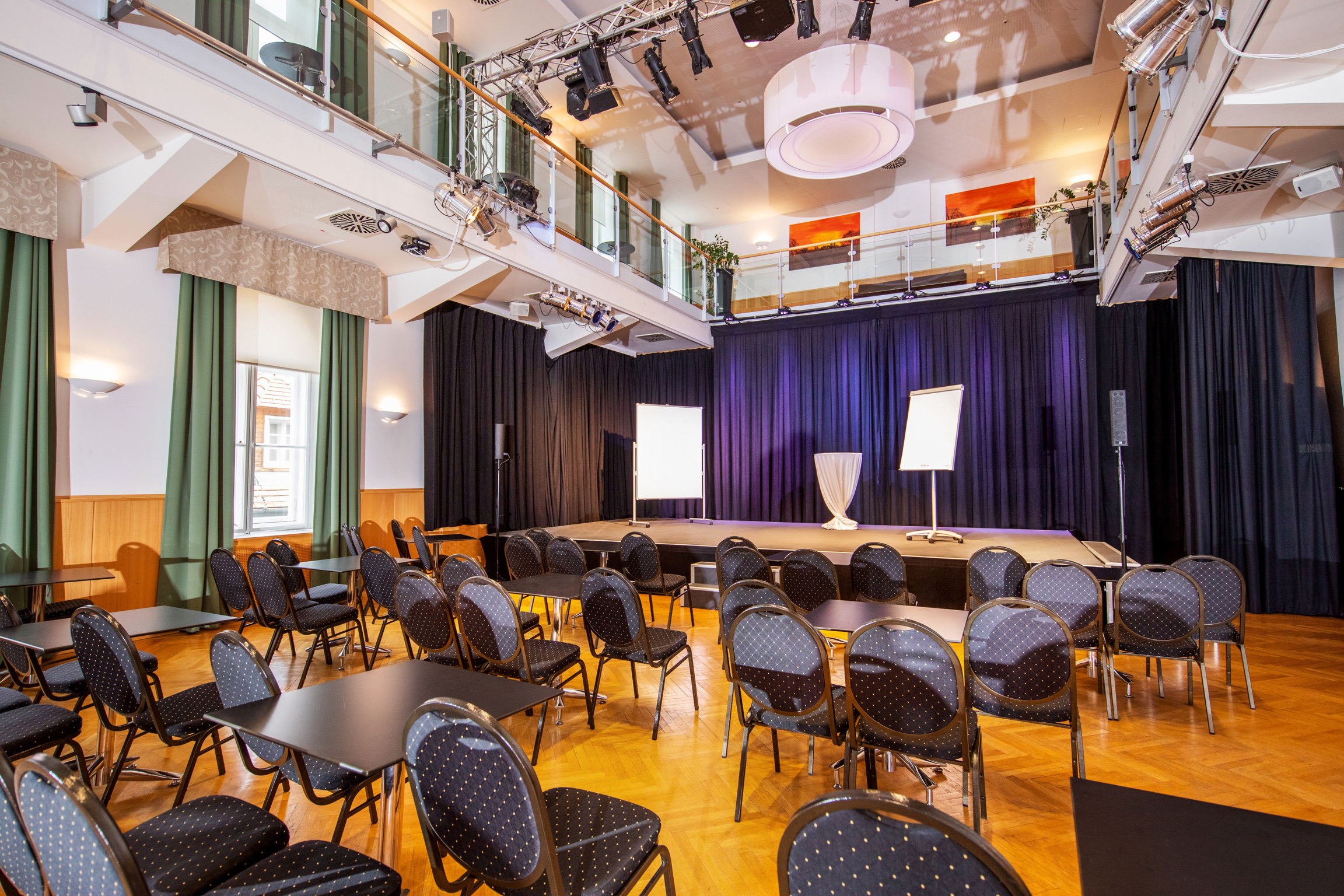 Ein Veranstaltungsraum mit einer kleinen Bühne, mehreren Stuhlreihen und Tischen, schwarzem Vorhang im Hintergrund, Bühnenleuchten an der Decke und einer oberen Galerie mit Glasgeländer.