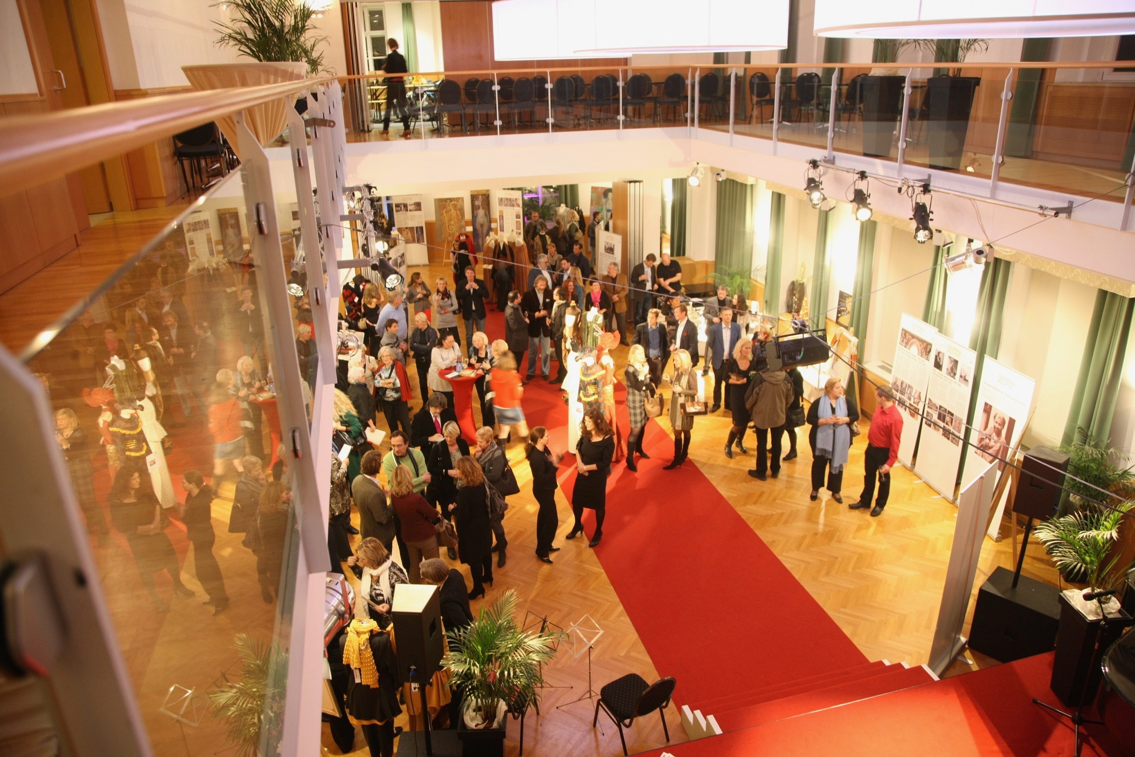 Blick auf eine Galerie mit Kunstwerken und Menschen bei einer Veranstaltung, im Vordergrund ein roter Teppich, im Hintergrund ein Balkon mit Sitzgelegenheiten.