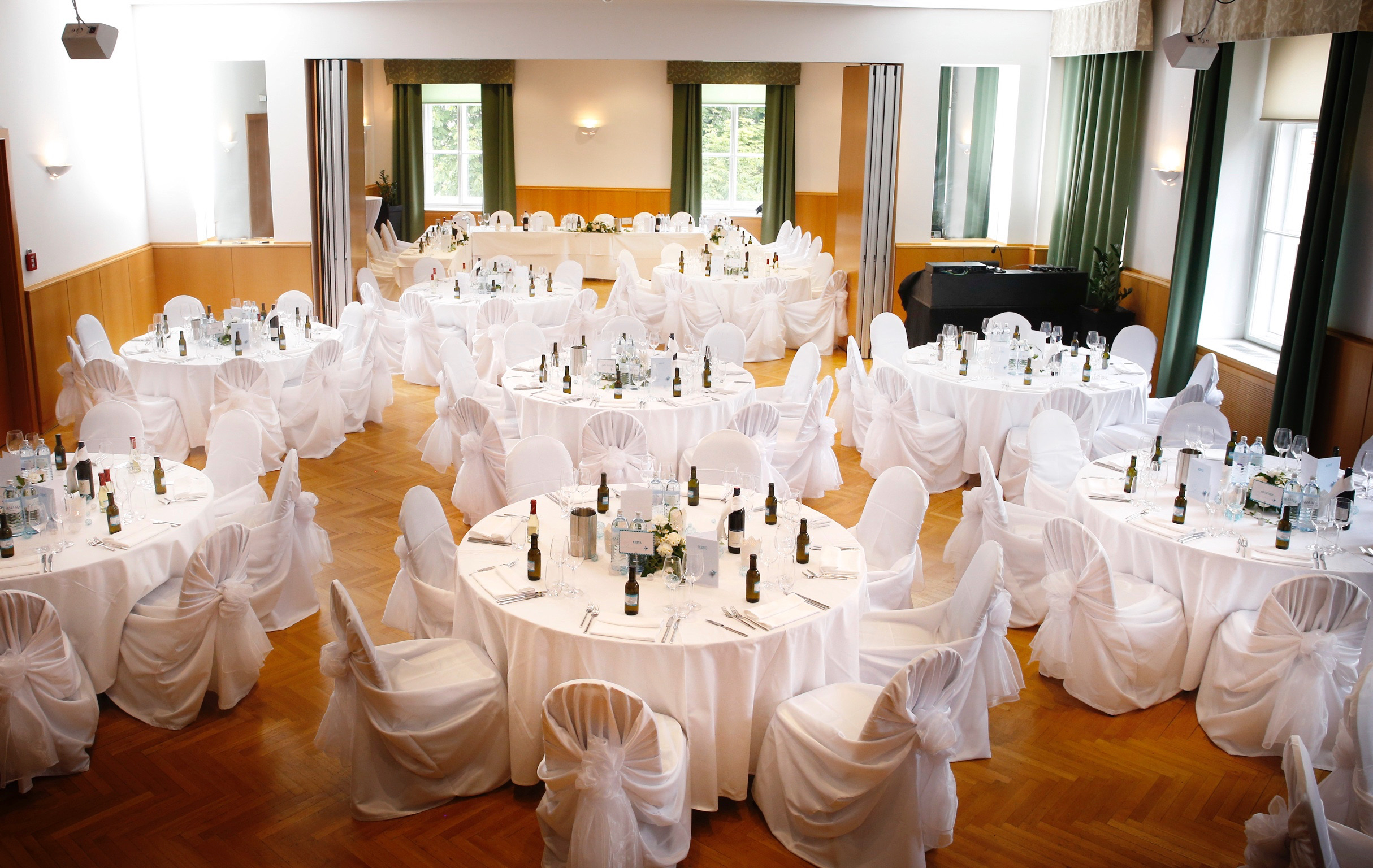 Eleganter Veranstaltungsraum mit runden Tischen, weiße Tischtücher und Stuhlhussen, festliche Dekorationen, Getränke und Besteck, große Fenster mit grünen Vorhängen.