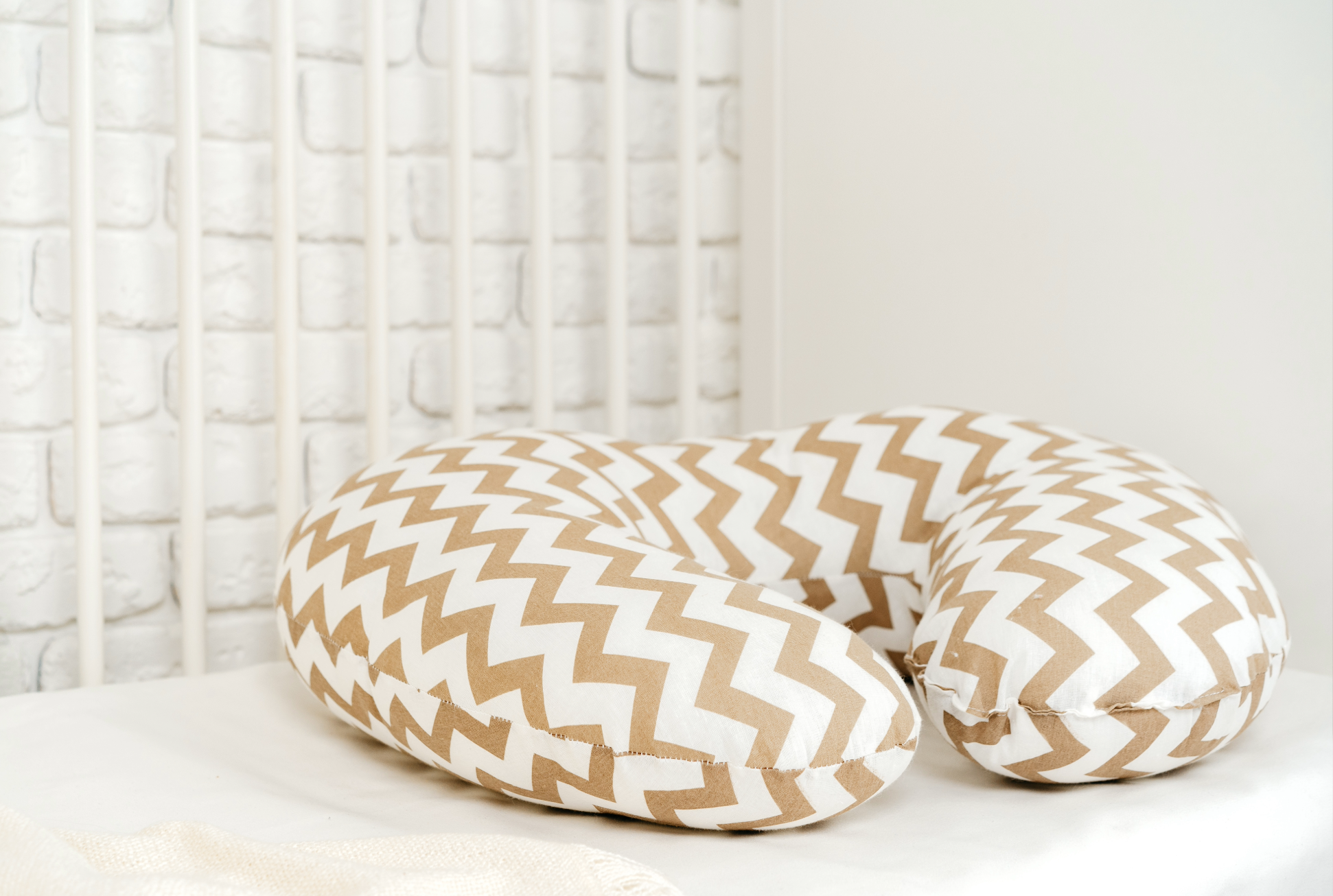 A nursing pillow with a brown and white zigzag pattern on a bed, with a white brick wall and white headboard in the background.