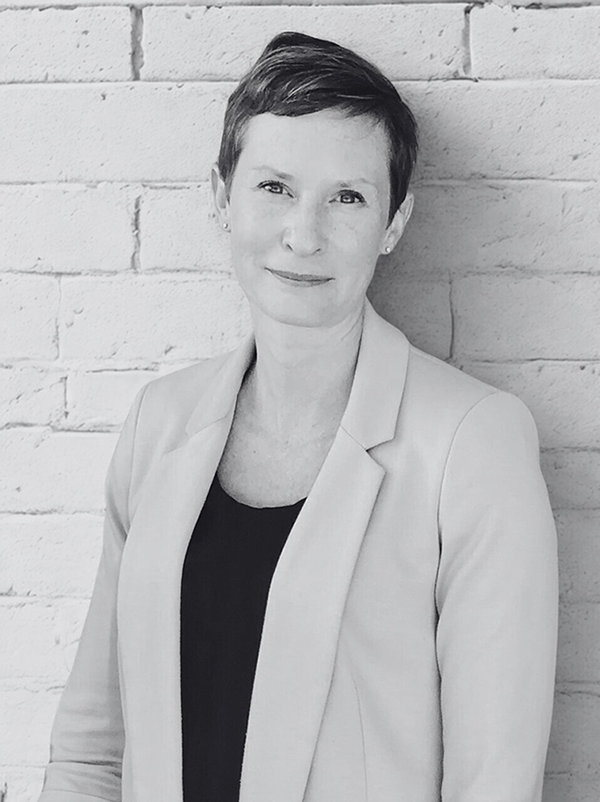 Black and white portrait of Jenn Taylor with short hair, wearing a blazer over a top, standing against a brick wall.