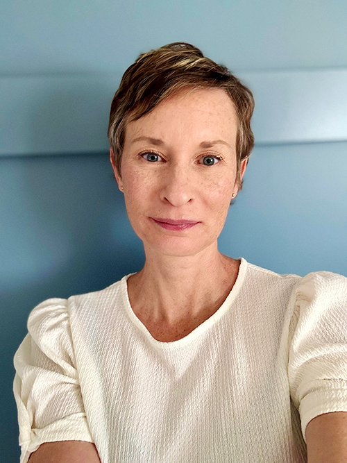 Close-up selfie of Jenn Taylor with short brown hair, wearing a white top, smiling softly, with a light blue wall in the background.