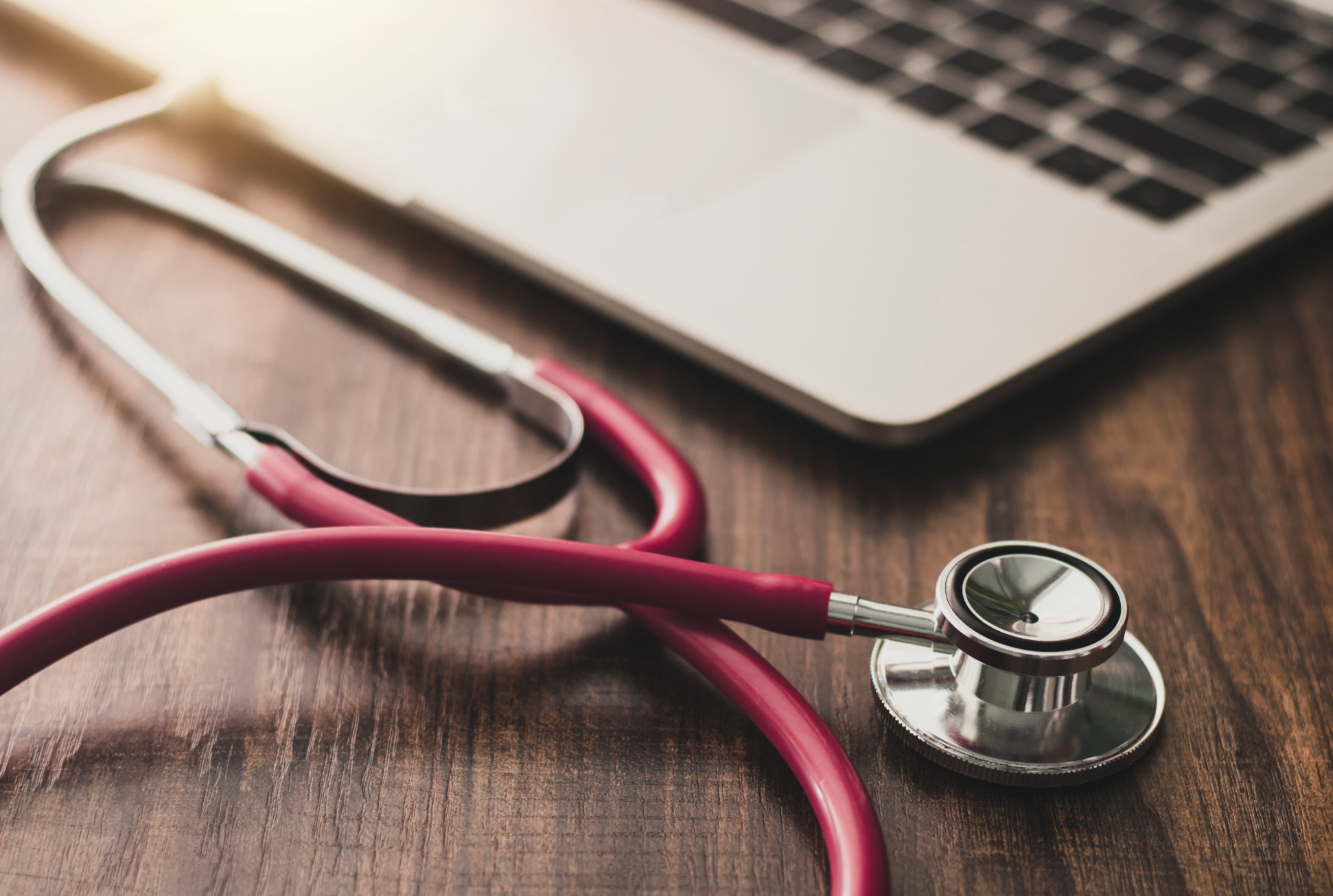 A stethoscope with pink tubing lying on a wooden desk next to a laptop.