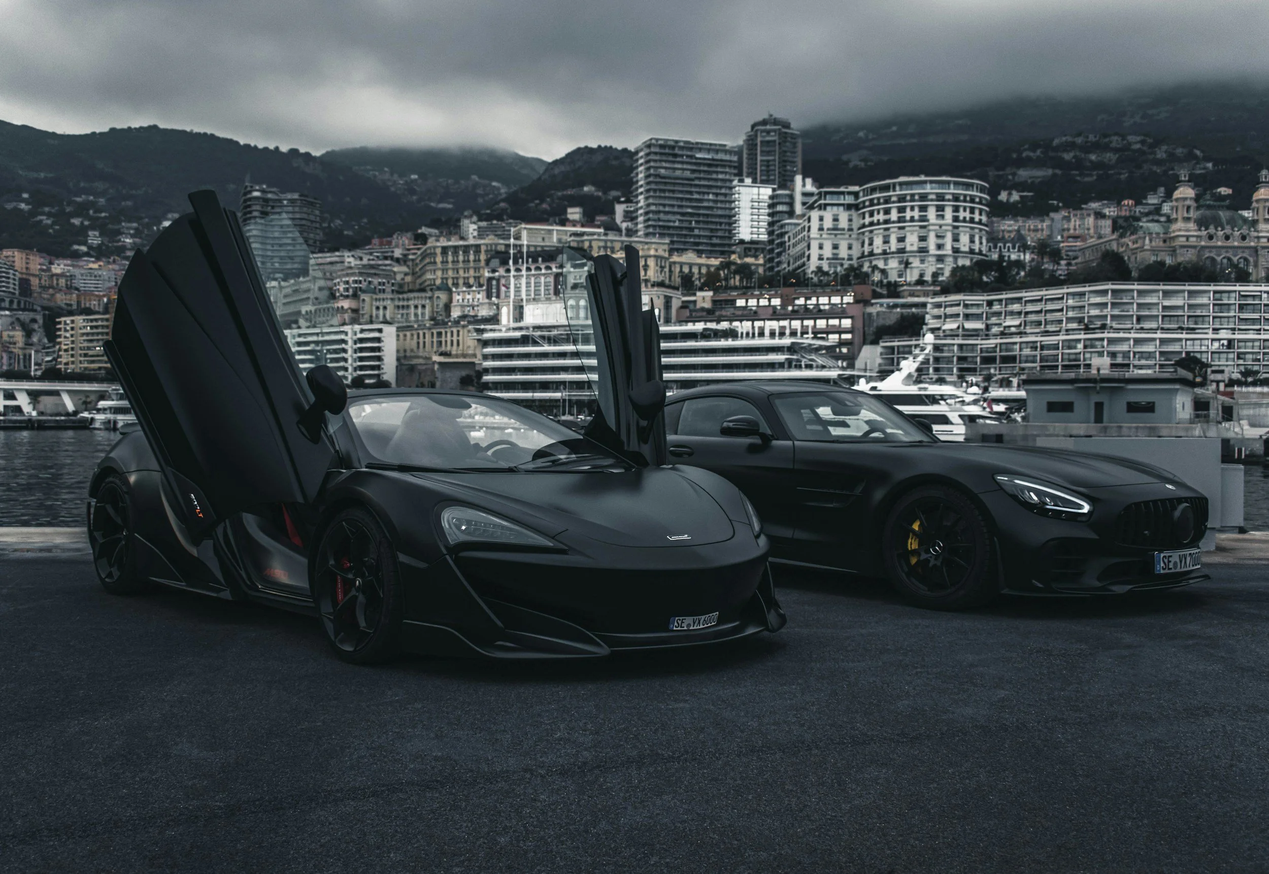 Two black luxury cars parked by a marina with city buildings and mountains in the background on a cloudy day.