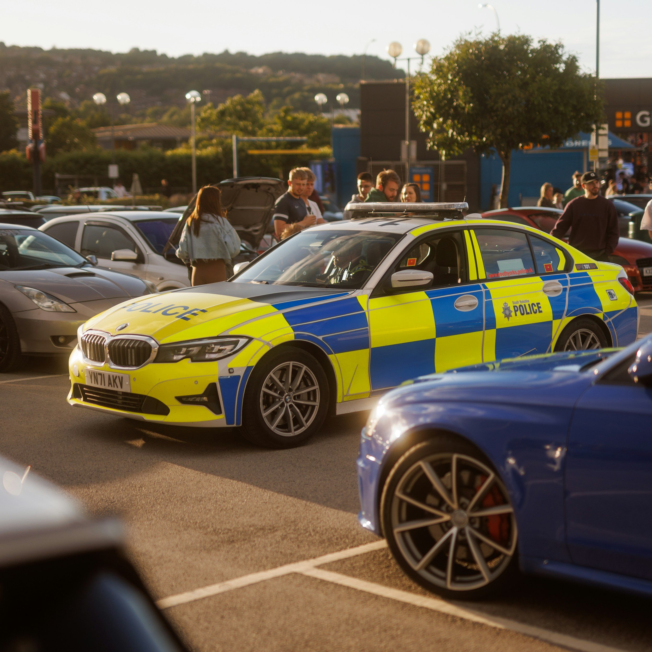 A police car parked in a busy parking lot with people around, some inspecting a vehicle with its hood open, during daylight