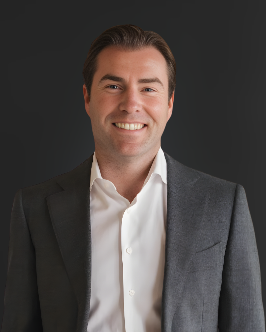 Headshot of a smiling man with brown hair in a white shirt and gray blazer against a dark background.