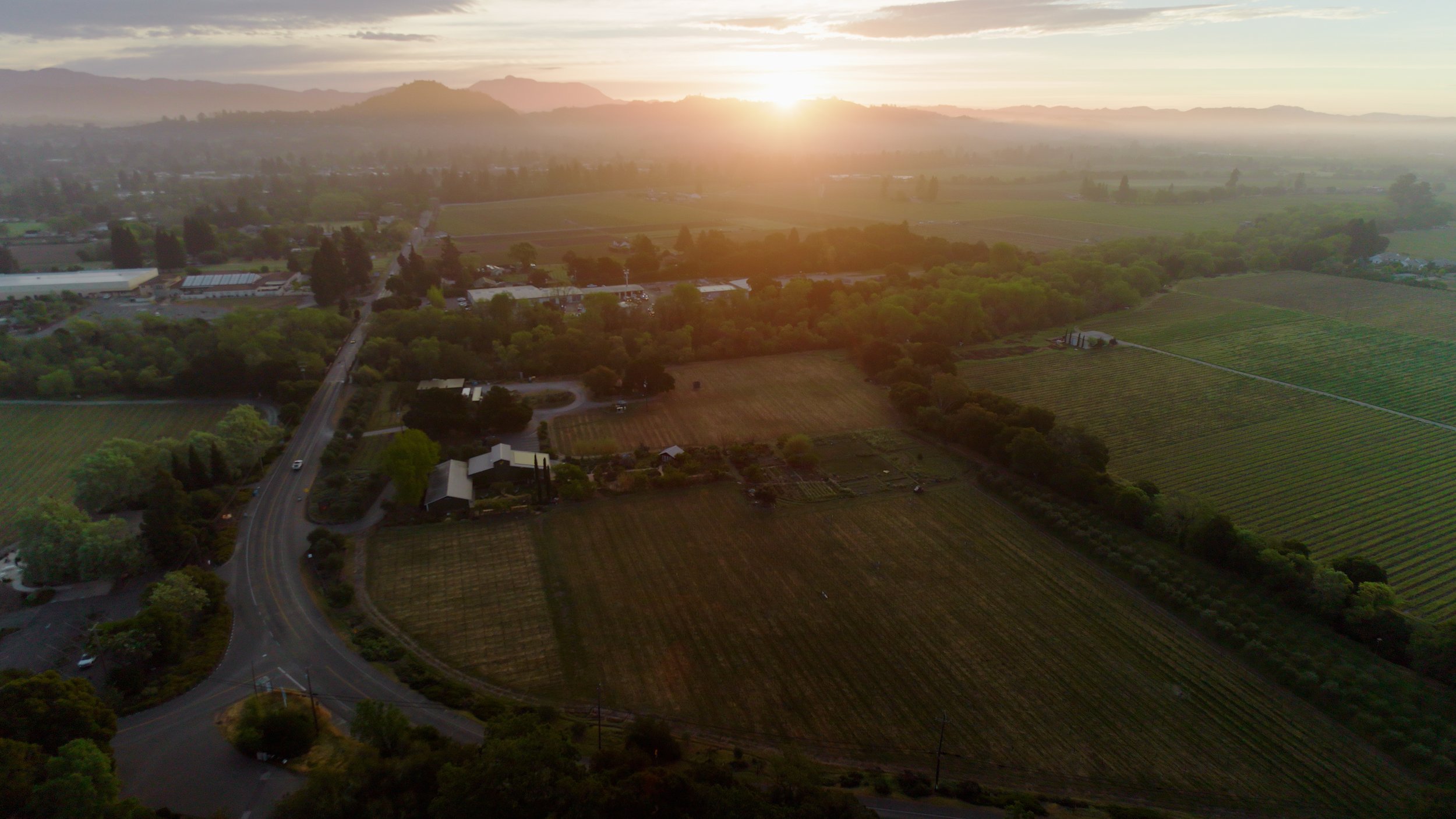 Aerial view of a rural landscape at sunset with fields, trees, roads, and distant mountains.
