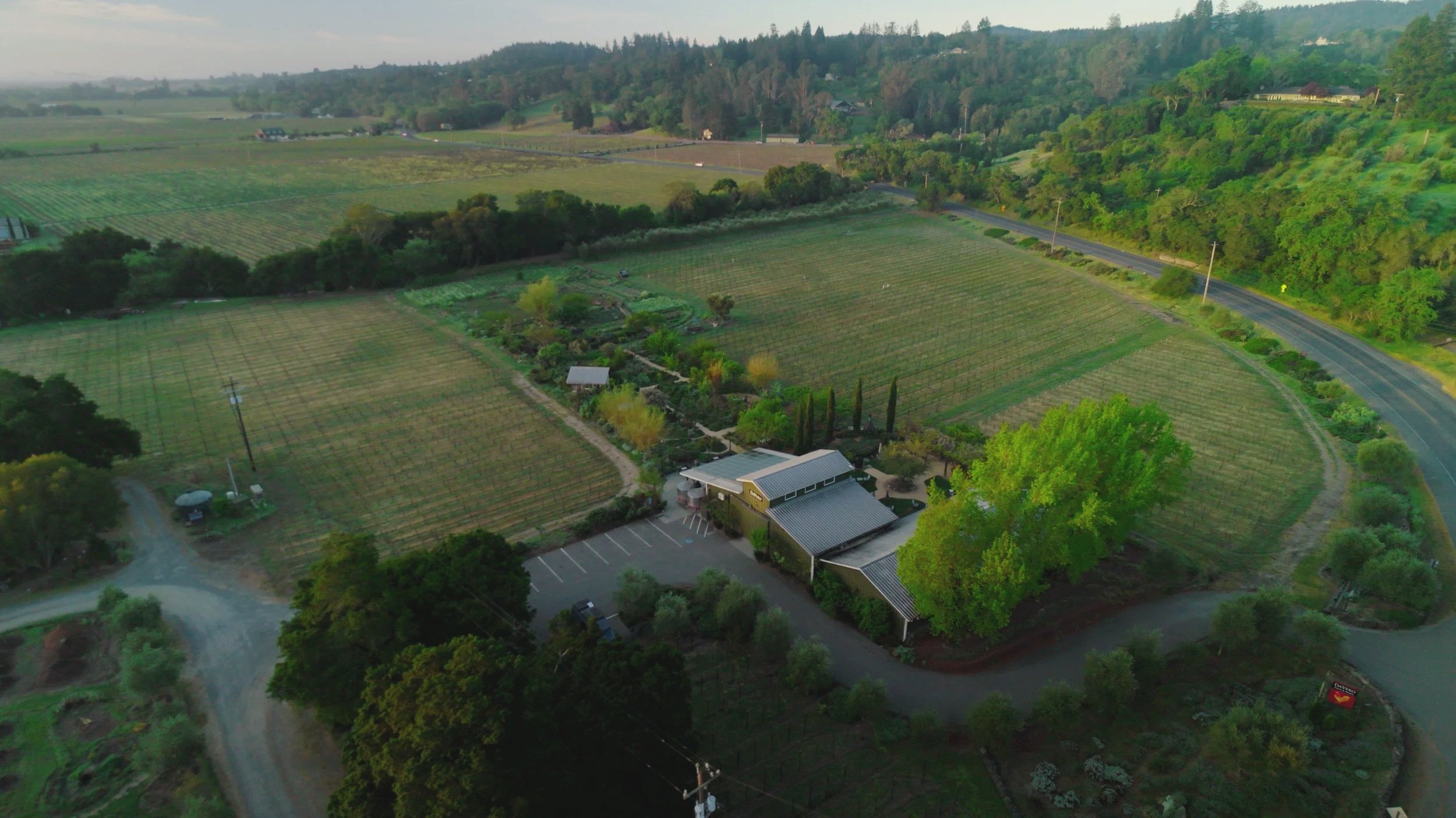 Aerial view of green farmland with fields, trees, a parking lot, and a building, surrounded by hills and a road.
