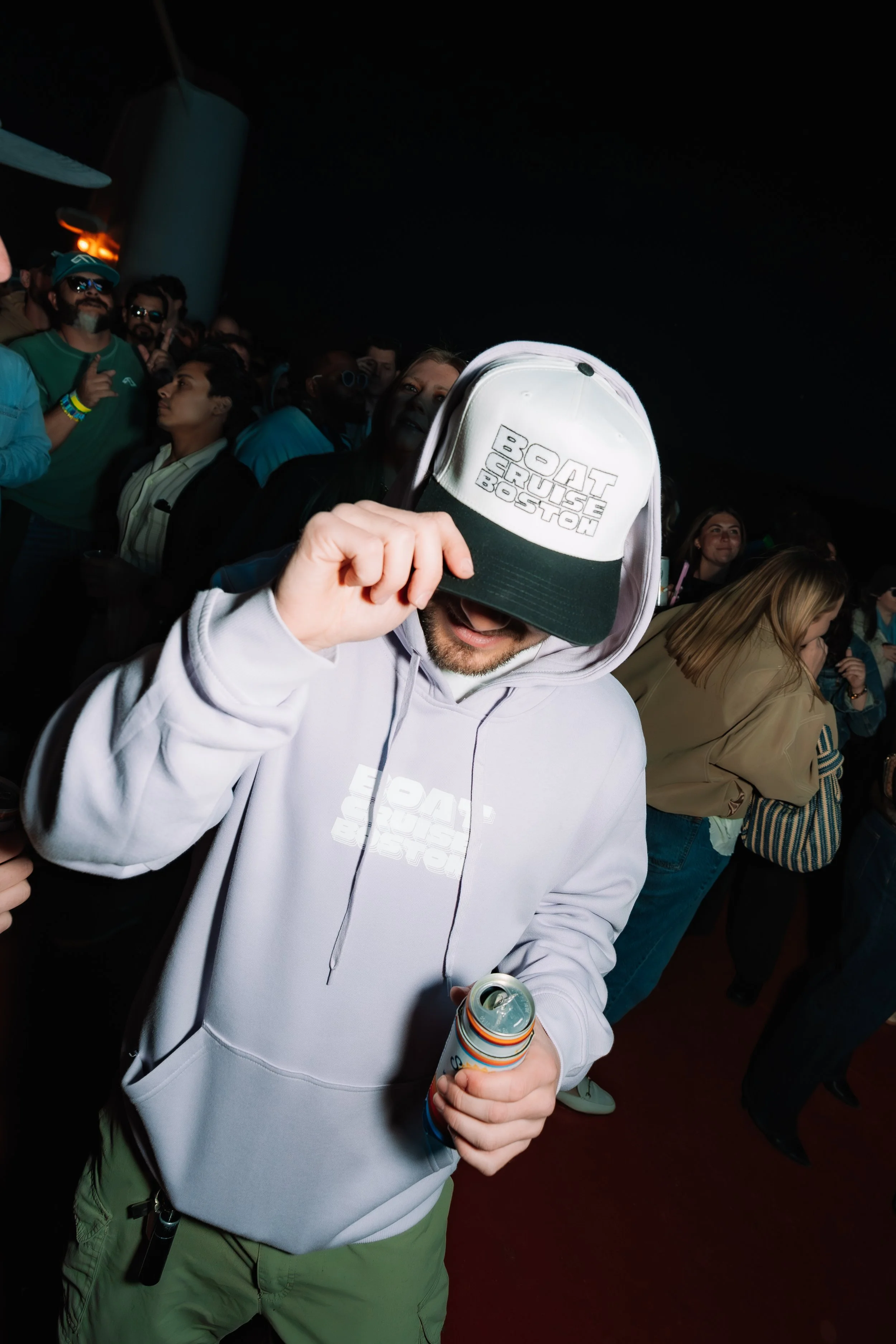 A man in a white hoodie and green pants holding a canned drink, partially hiding his face behind a white and black trucker hat reading 'BOAT TOUR BOSTON', in a crowded indoor setting.