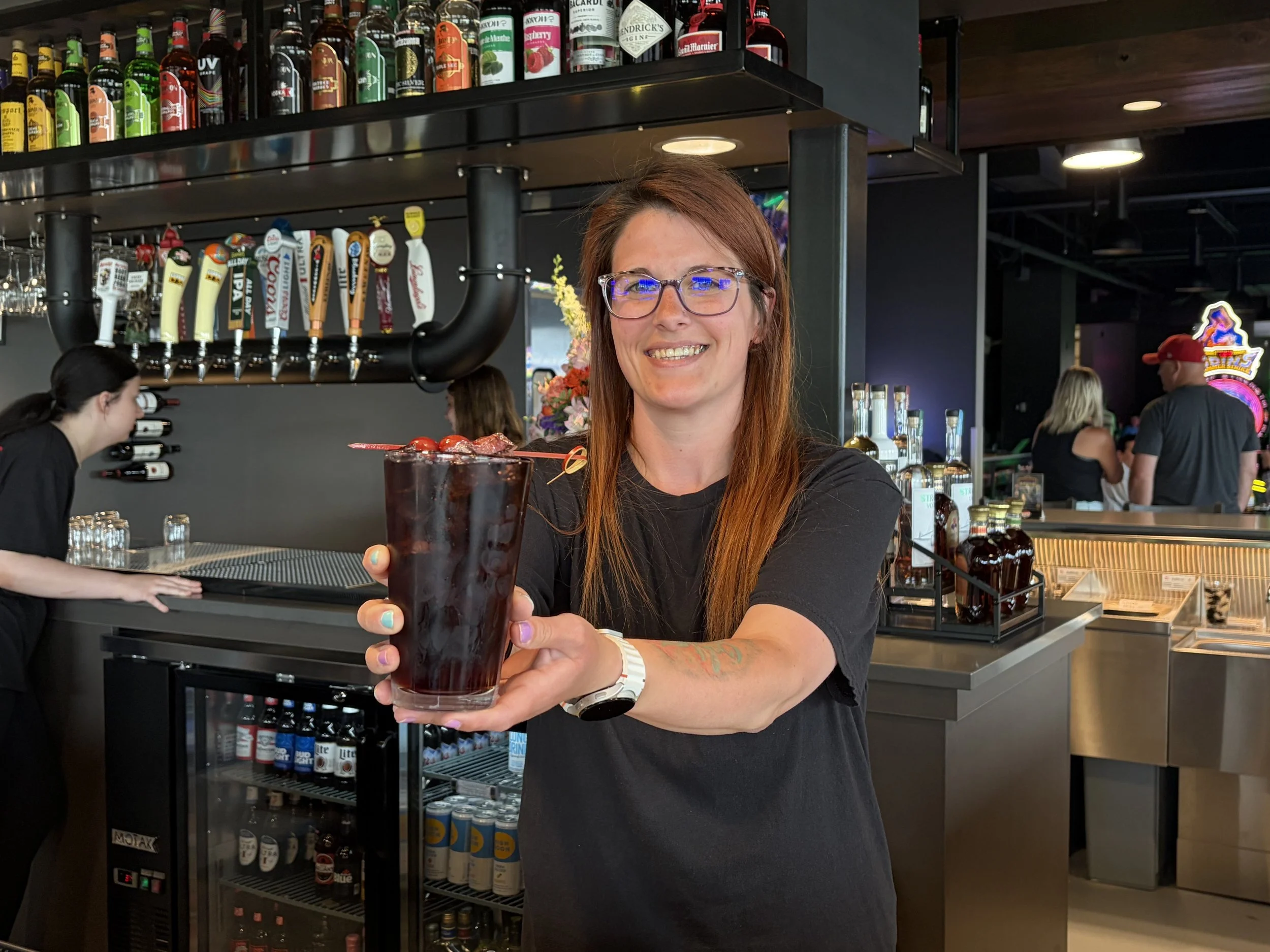 A woman with glasses and red hair smiling and holding a large glass of dark-colored cocktail inside a bar or restaurant. The background shows a bar counter with various bottles of alcohol, beer taps, and other patrons.