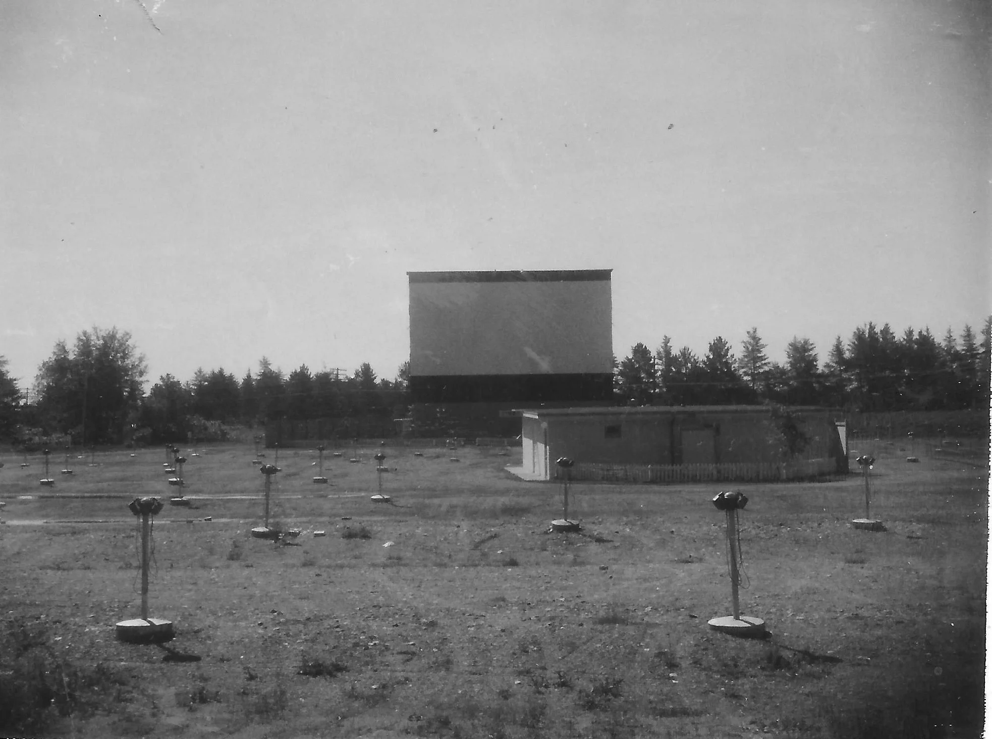 Black and white photograph from 1962 of a drive-in movie theater property with speakers spaced evenly through the lot. In the background, there is a large rectangular movie screen and a line of trees under a mostly clear sky.