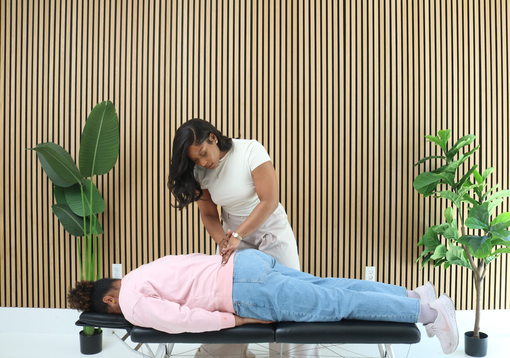 A woman in a white shirt assisting a person lying face down on a massage table, in front of a wood-paneled wall with two large green plants on either side.