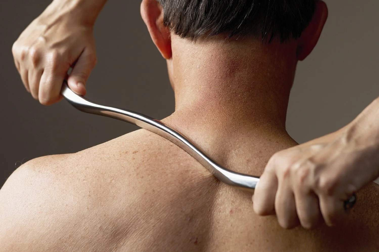 A person performing Graston Technique with a metal massage tool on the back of a person's neck and shoulder area.