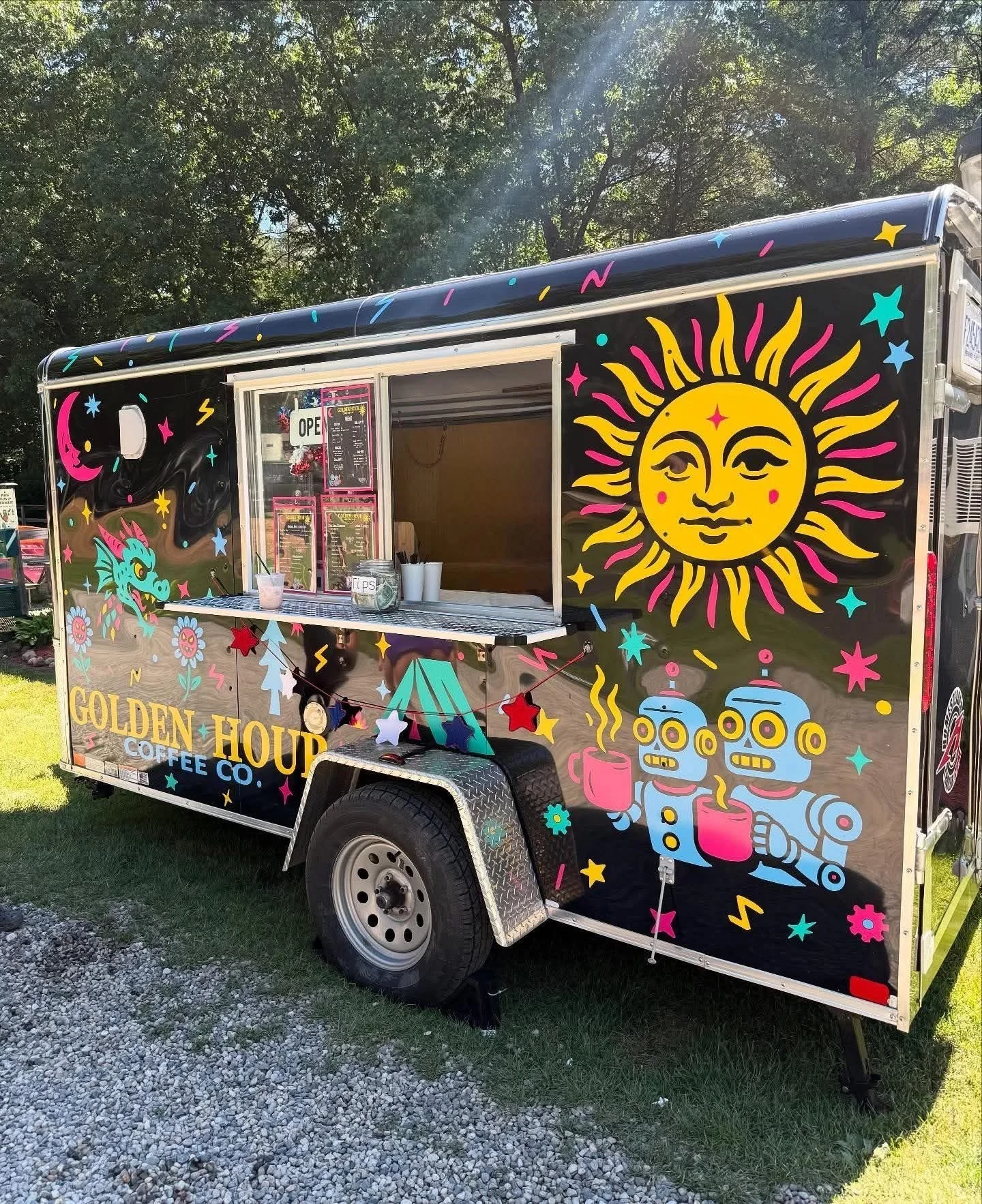 A colorful food truck with a dark blue background, decorated with vibrant celestial and fantasy-themed illustrations, including a prominent smiling sun with wavy rays, a smiling moon, a dragon, and whimsical stars and clouds. The truck has the words "Golden Hour Coffee Co." written on it.