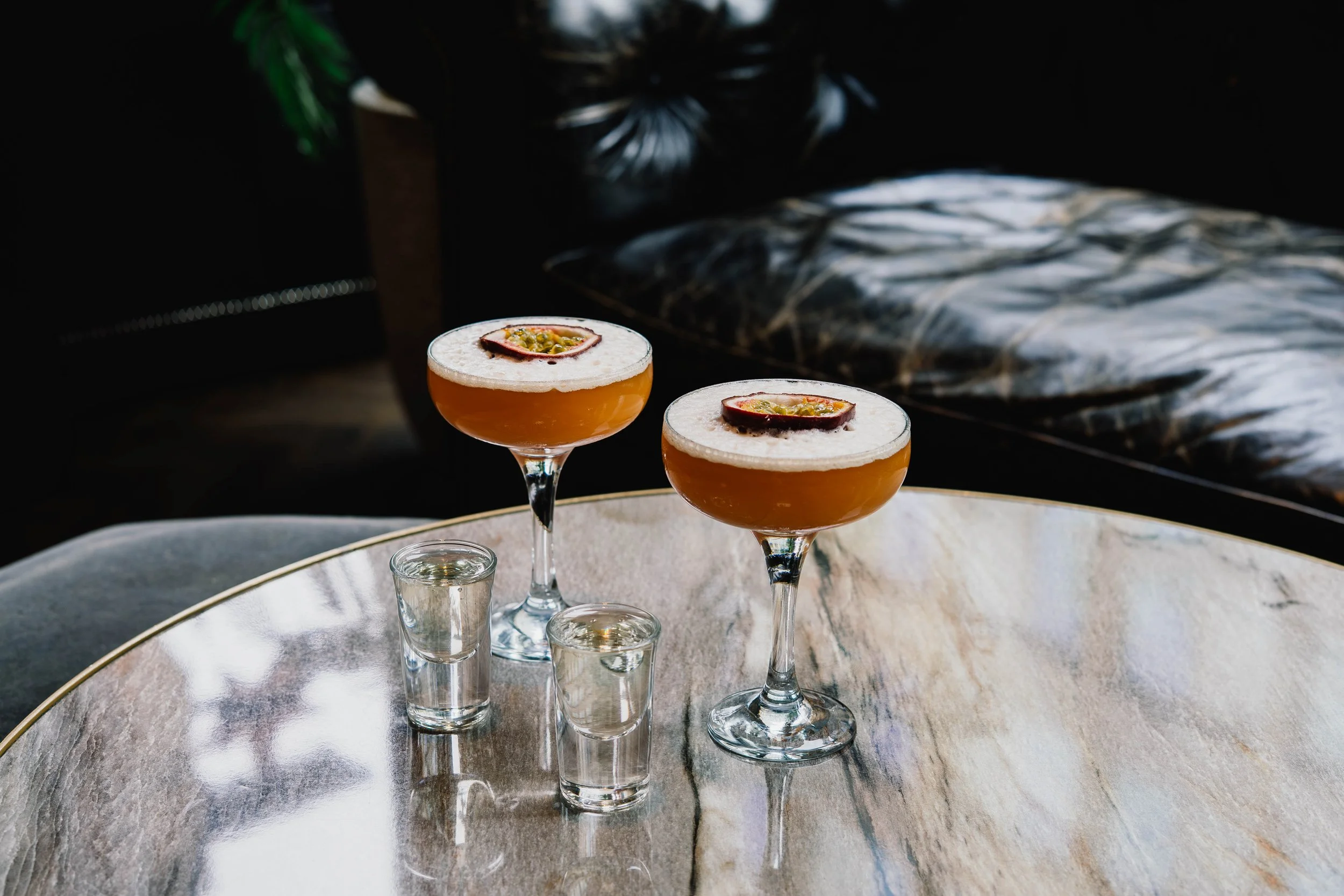 Two Passionfruit cocktails on a table with three empty shot glasses in front of them, a dark background with a blurred patterned surface, and a large silver bowl surrounding the table.