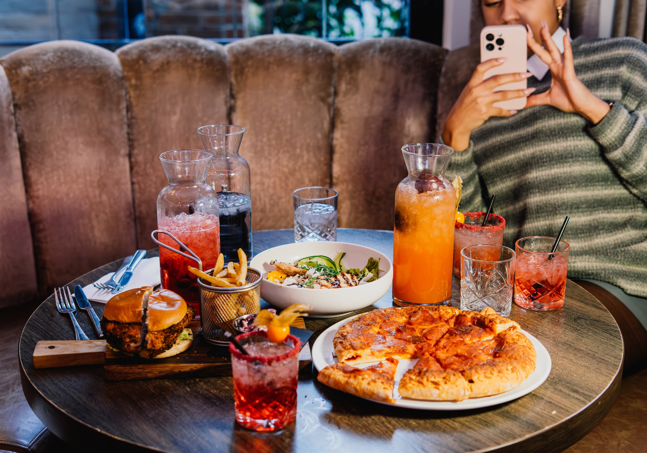 A table full of various food and drinks, including pizza, pizza burger, pasta, salads, and multiple colorful beverages, with a woman taking a photo of the meal with her smartphone.