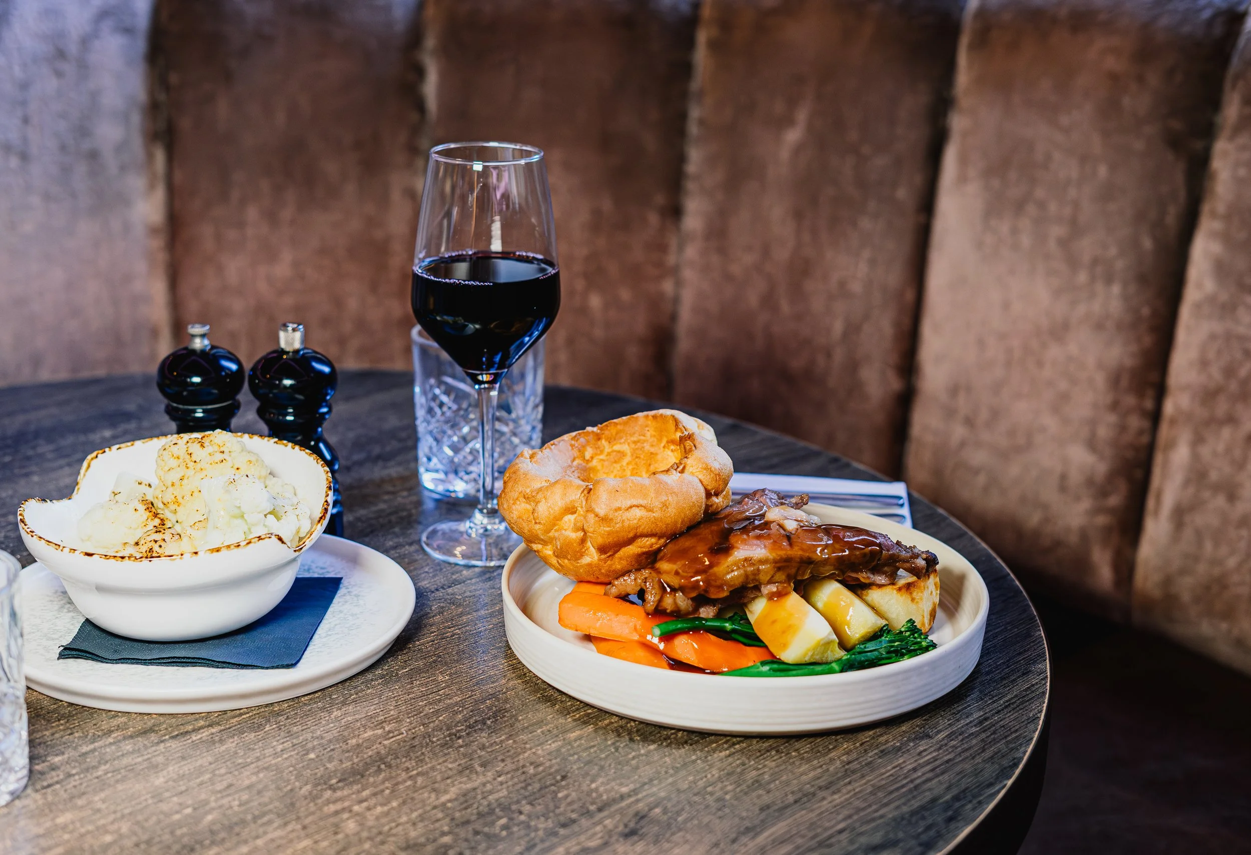 A dining table with a glass of red wine, a bowl of mashed potatoes, a bread roll, small bottles of condiments, and a cooked meal with vegetables and meat. The background features wooden panels.