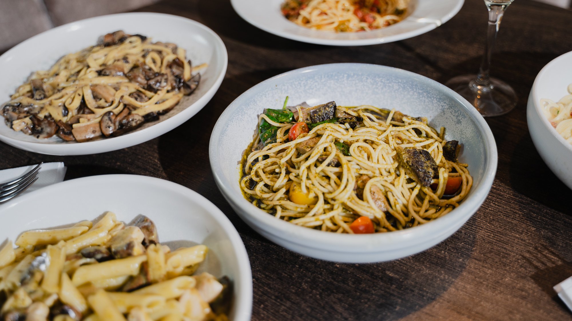Various plates of pasta dishes on a dark wooden table, including a bowl of spaghetti with vegetables and a plate of mushroom pasta.