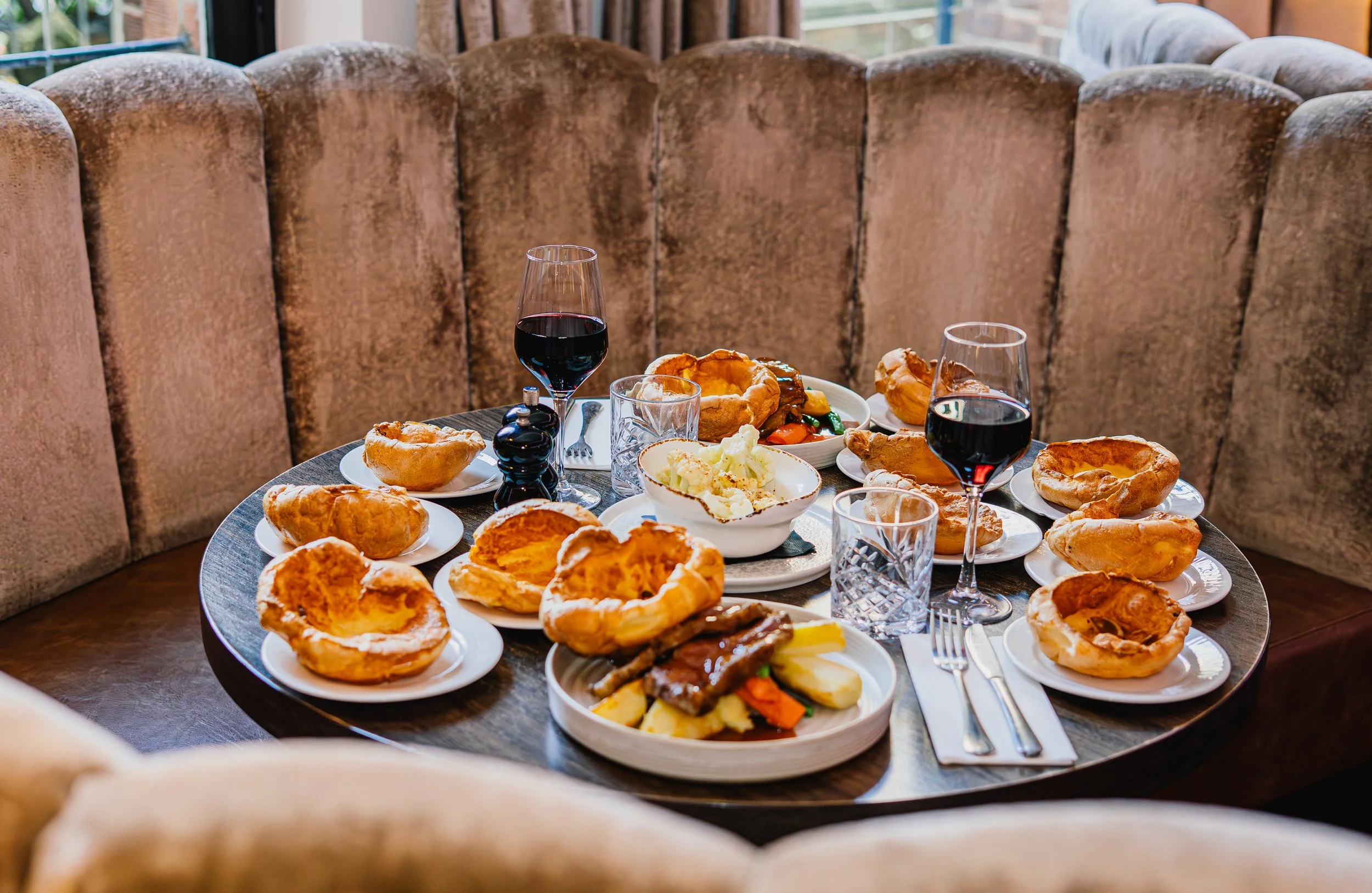 A round table filled with plates of half-eaten pie and food, two glasses of red wine, and a pepper grinder, set in a cozy dining area with a cushioned booth in the background.