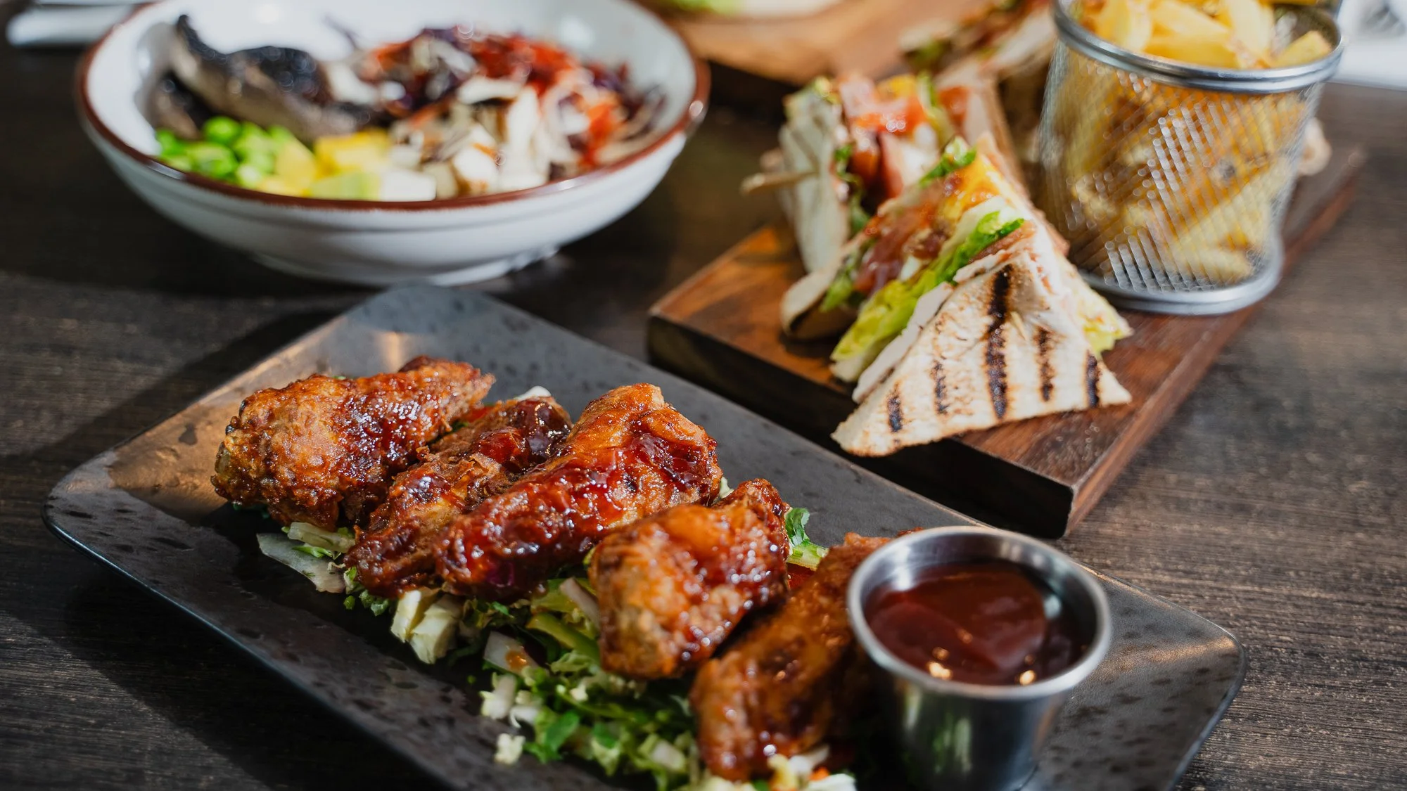 Close-up of a serving tray with barbecued chicken drumsticks topped with sauce, lettuce, and shredded vegetables, accompanied by a small cup of barbecue sauce. In the background, there is a wooden board with grilled sandwiches filled with vegetables 