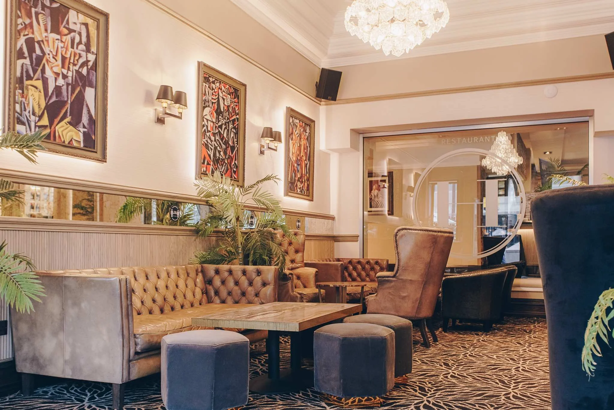 A cozy hotel lobby with leather tufted sofas, armchairs, a large chandelier, framed artwork, plants, and a mirror reflecting warm lighting.