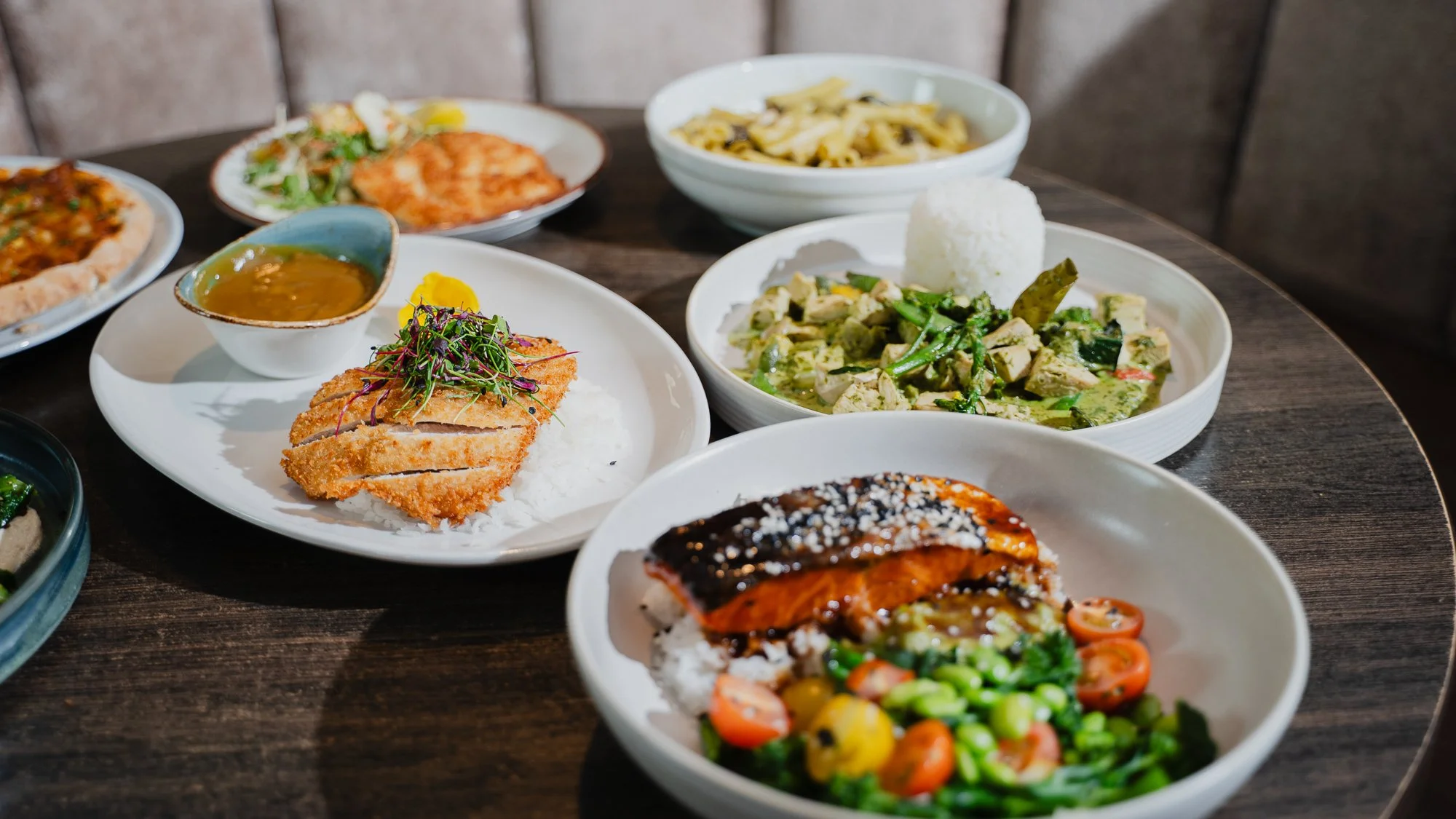 Assorted Asian dishes on white plates and bowls, including rice, curry, grilled fish, salad, and noodle dishes, arranged on a wooden table.