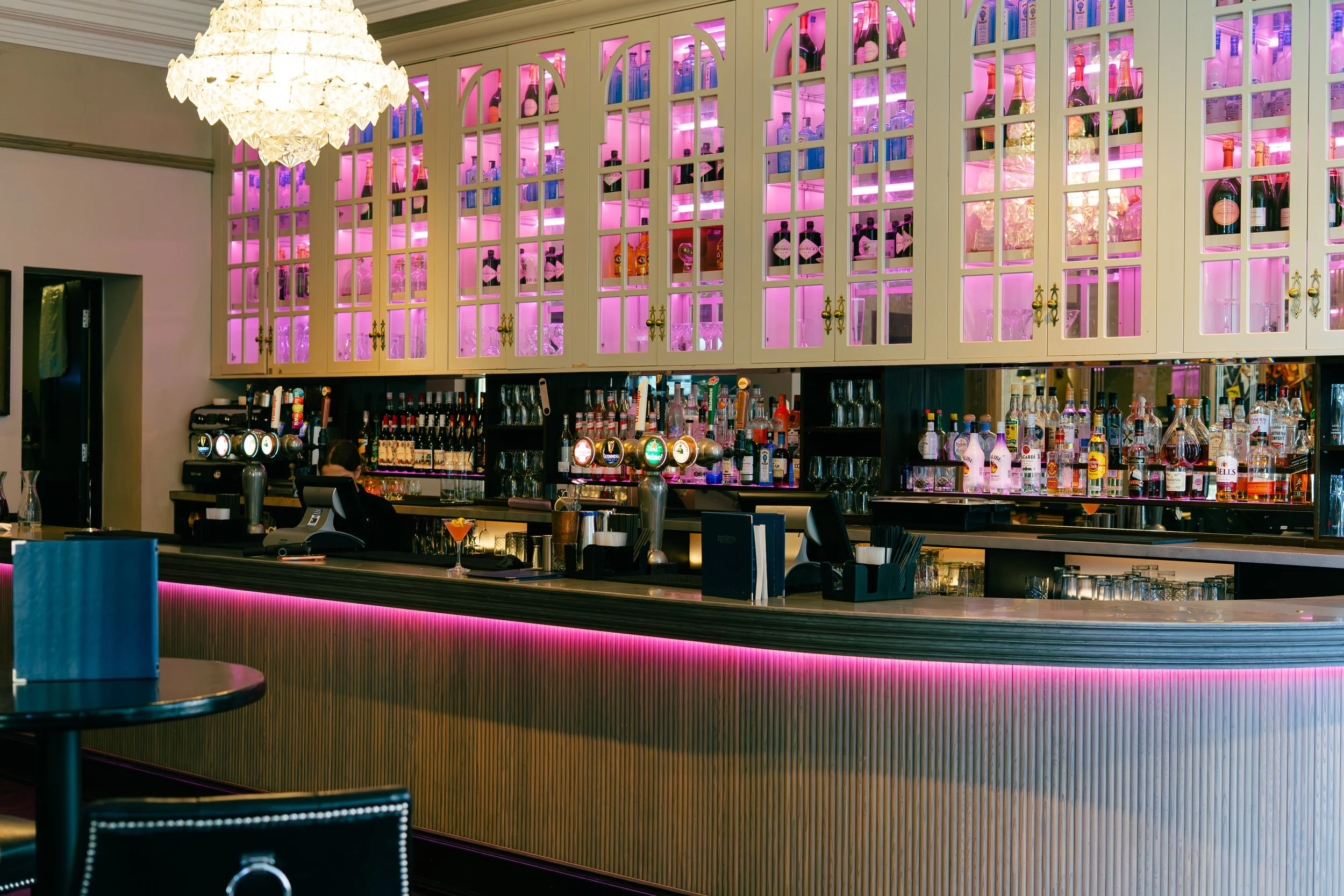 Bar with glass shelves illuminated in pink holding bottles of alcohol, beneath a curved bar counter with a pink light strip and a decorative chandelier overhead.