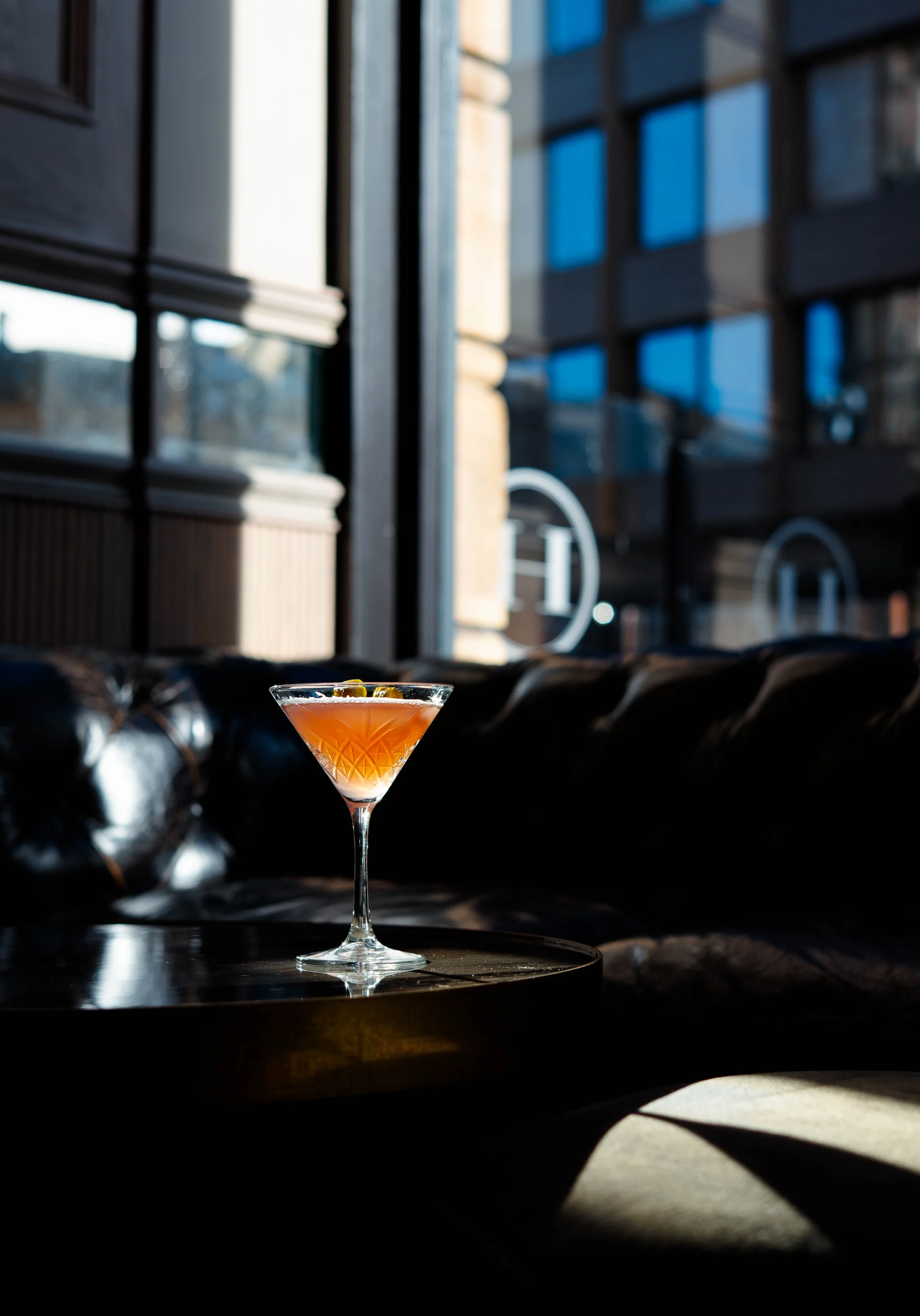 Close-up of a pink cocktail in a martini glass with lemon peel garnish, set on a dark table against a blurred background of modern buildings and large windows.