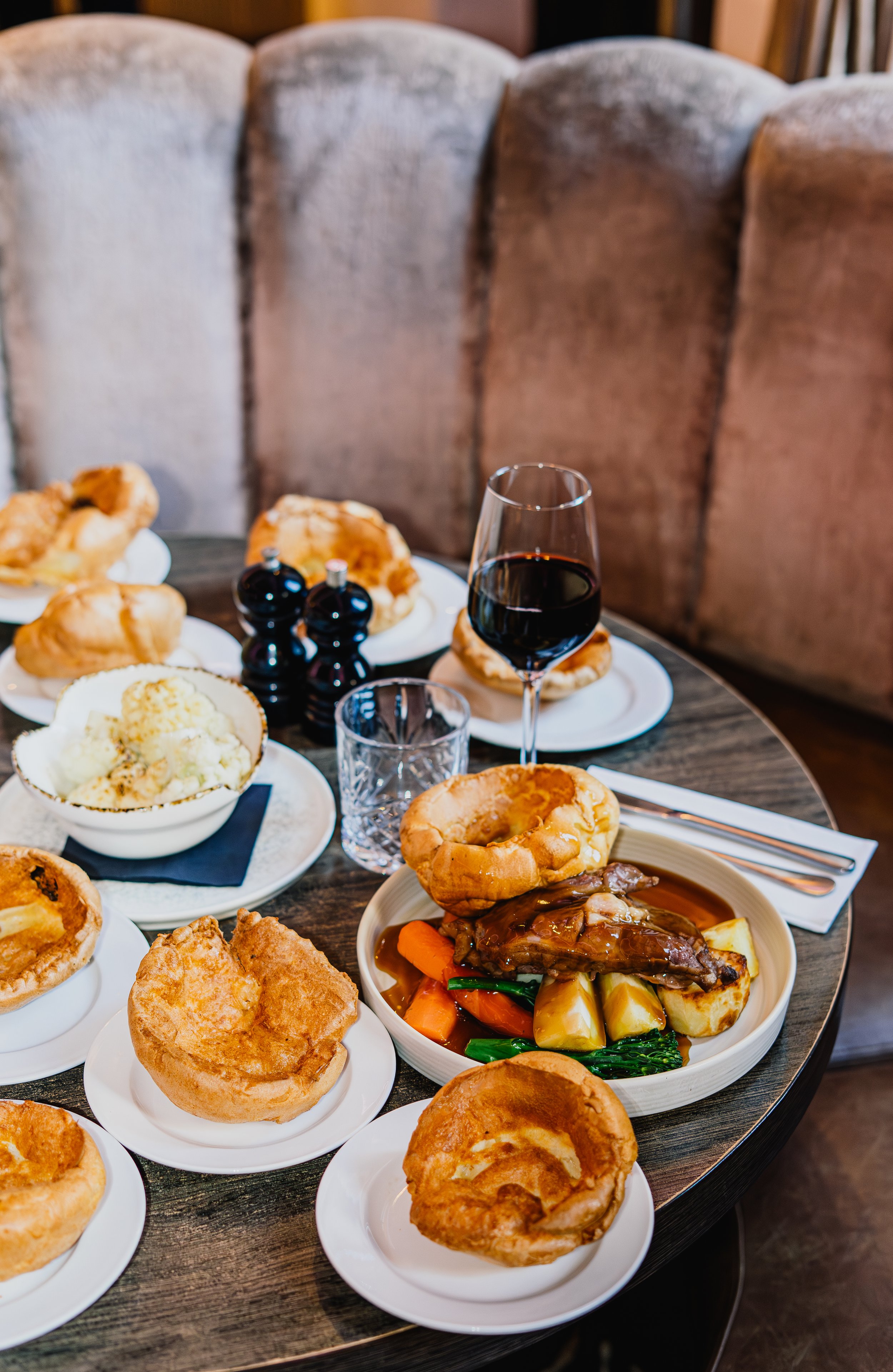 A table filled with various plates of food, including pies, roasted vegetables and meat, a glass of red wine, and salt and pepper shakers, set in a restaurant.