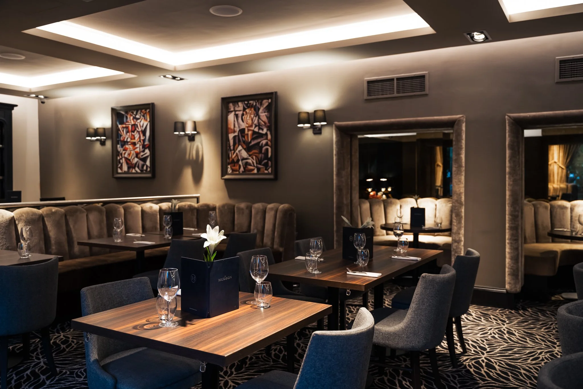 A cozy restaurant dining area with arranged tables, wine glasses, and a black box with a white flower on each table. The room features neutral tones, modern artwork on the walls, and warm ambient lighting.