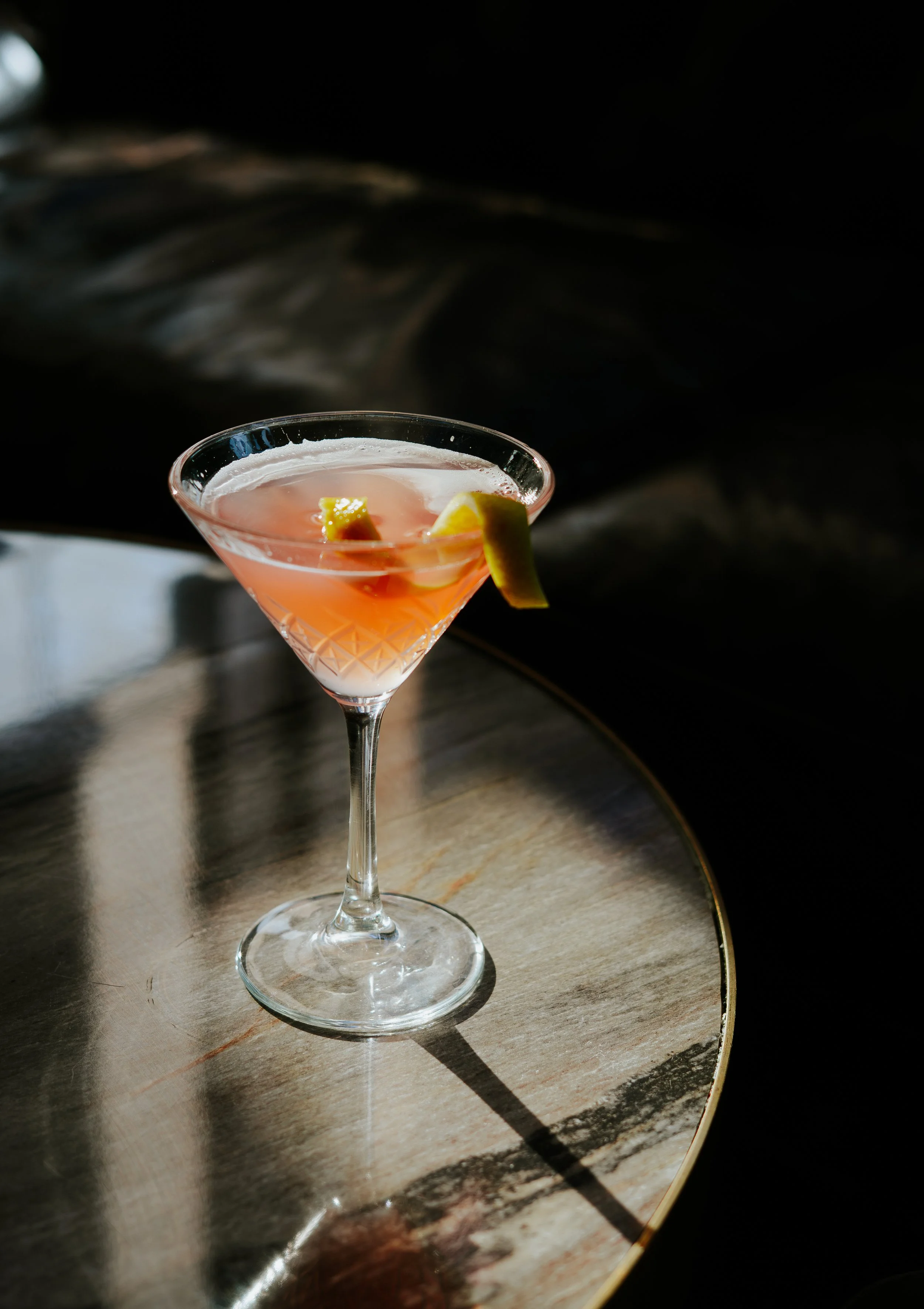 A pink cocktail in a martini glass garnished with lemon wedges, placed on a wooden table with a reflective surface.