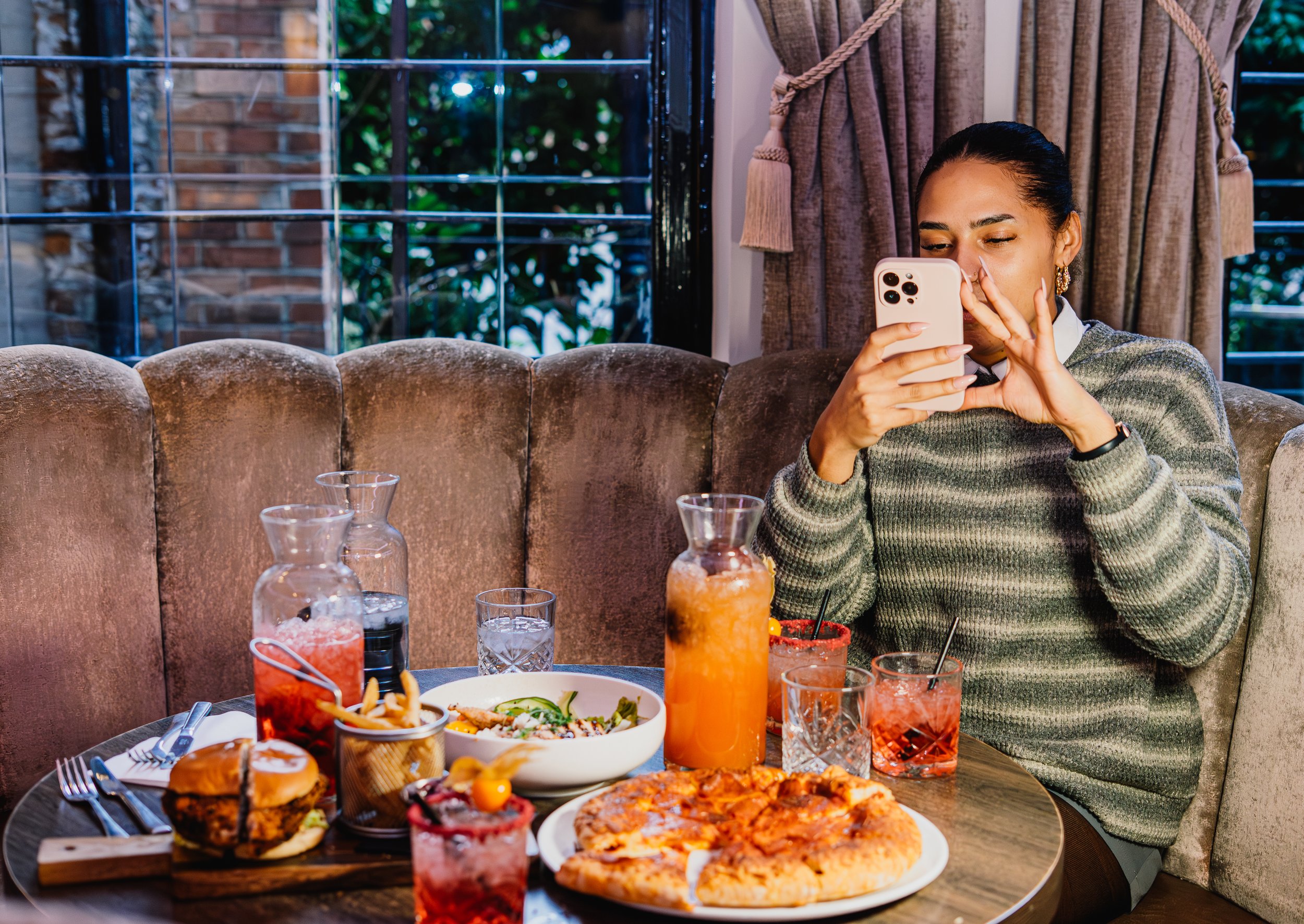 A woman sitting at a restaurant table, looking at her phone, with a variety of food and drinks in front of her, inside a cozy dining area with curtains and windows showing greenery outside.